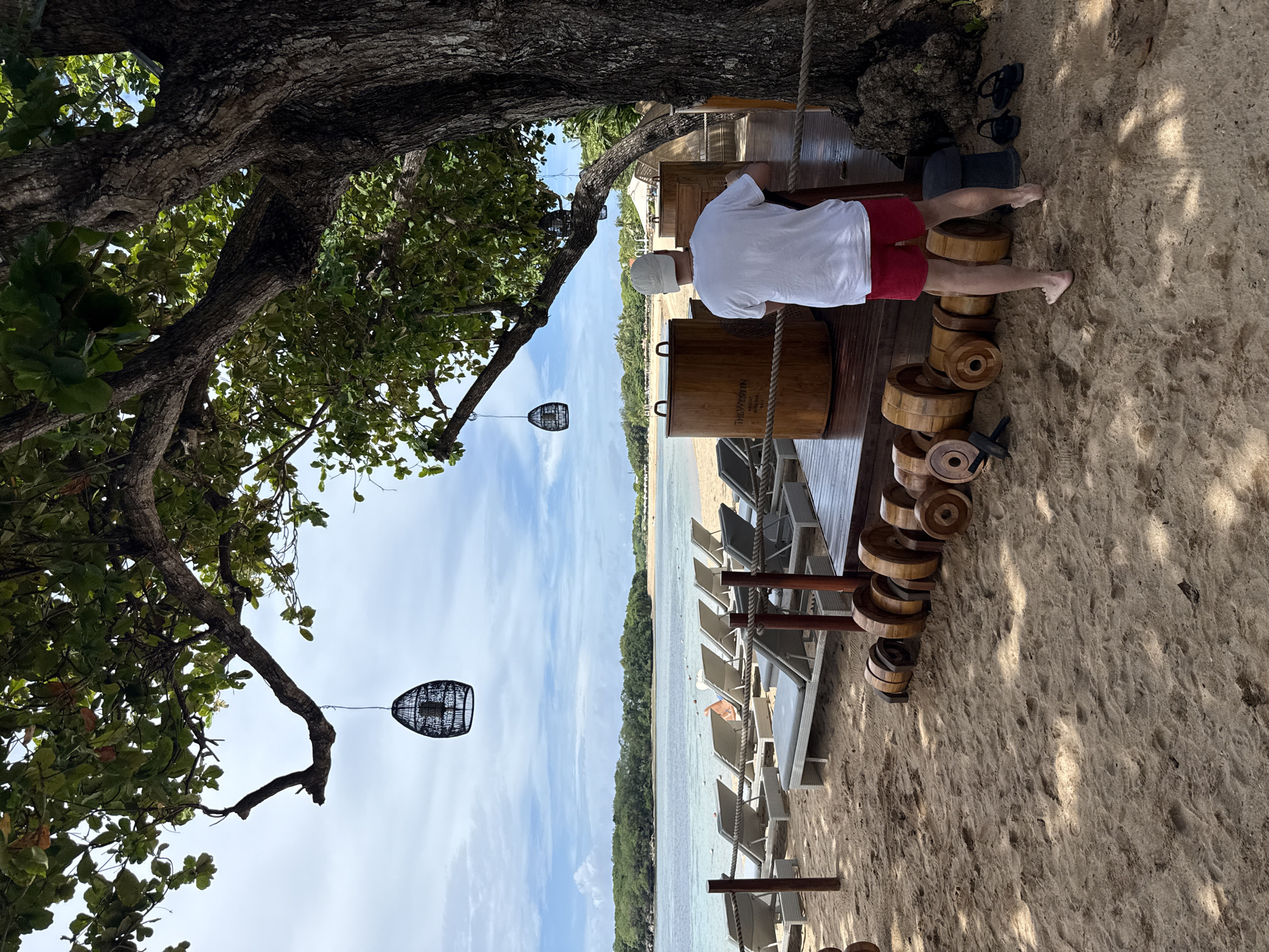 The image depicts an outdoor scene at the beach in Kuta Selatan, Indonesia. A man wearing a white shirt, red shorts, and a white cap is leaning against a wooden structure adjacent to several large, wooden barrel-shaped containers. He is barefoot. Beside the man, on the sand, are a pair of sandals. To the left, a tree with green leaves and decorative lanterns is visible. In the background, there is a beach with numerous lounge chairs, the ocean, and a line of trees along the coastline. The sky is partly cloudy. The angle of the photograph suggests it may have been taken from a slightly elevated position.