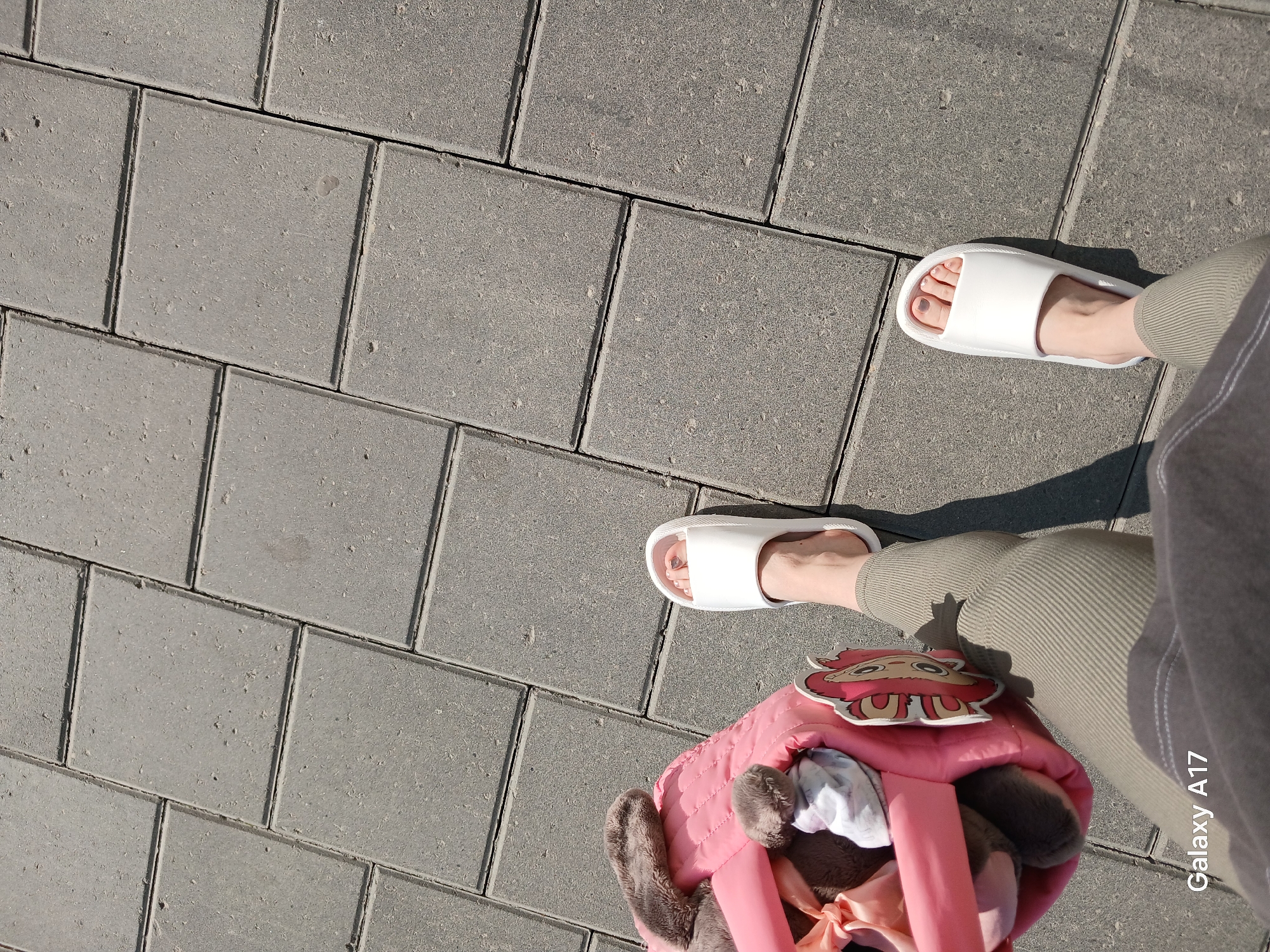 The image shows a street scene in Yekaterinburg, Russia. The surface is paved with gray, square paving stones. A person is standing on the pavement, wearing white slides on both feet, olive green pants, and a gray top. A pink bag with a cartoon character attached is next to the person's left leg. The bag contains clothes and stuffed toys. The image has a "Galaxy A17" watermark.