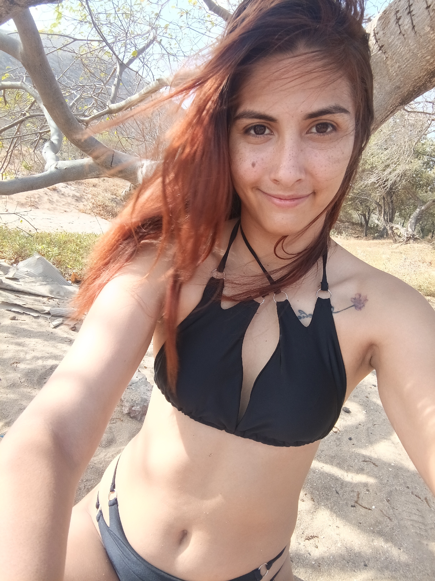 A woman with reddish-brown hair is taking a selfie in what appears to be a dry, wooded area. She's wearing a black bikini. The background shows sparse vegetation, including bare trees and dry ground. The location is identified as Pedro González, Venezuela. A tree branch extends from the upper left of the frame. The woman has freckles and a tattoo on her shoulder. The ground appears to be sandy. The lighting suggests it is daytime.