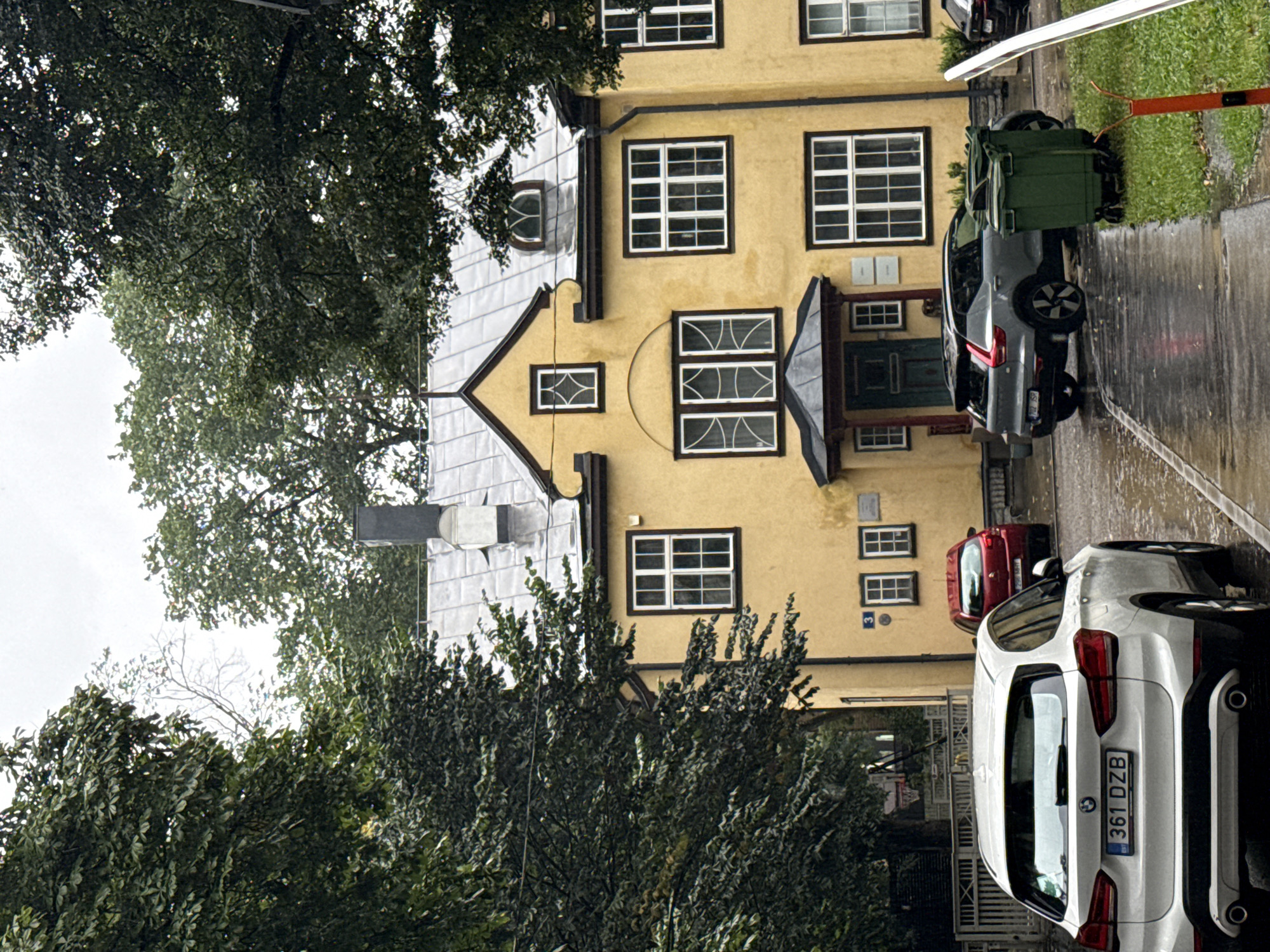 The image depicts a residential building facade painted a bright yellow, with a grey, sloped metal roof and several windows. The building is partially obscured by lush green trees on the left and right. In the foreground, a white BMW SUV is parked on a wet paved surface, which appears to be a street or driveway. Its license plate reads "361 DZB". To the right of the BMW, a grey Volvo SUV is parked on the same wet surface, with two green trash bins situated on a patch of grass next to it. The ground is reflective, indicating it has recently rained. In the background, a red car is parked further down the street. The overall scene is calm and appears to be a residential area in Tallinn, Estonia.