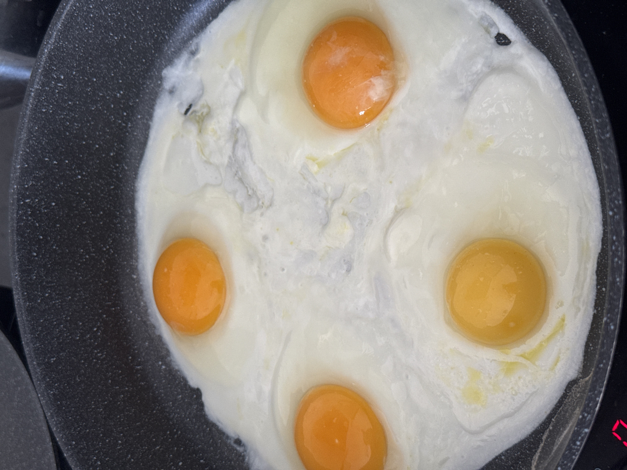A frying pan contains four fried eggs. The eggs have bright orange yolks and white, slightly opaque egg whites that have spread to the edges of the pan. The frying pan is dark gray with a speckled texture and is on a black stovetop. There is a small black speck in the white of the egg on the bottom right. A red digital display is visible at the bottom left of the image. The image is taken from a top-down perspective. This image does not depict a location in Lyubertsy, Russia.