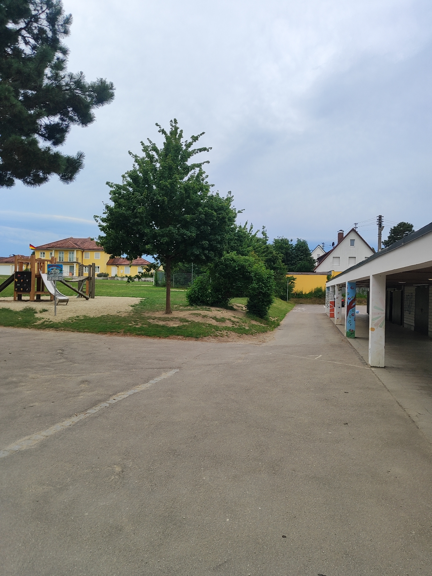 The image depicts an outdoor scene in the city of Diedorf, Germany, likely an educational facility or recreational area. In the foreground, a grey asphalt surface with white line markings extends into the mid-ground. To the left, a sandpit with a wooden playground structure featuring a slide and climbing wall is visible. Behind the playground, a large, yellow building with a red-tiled roof stands, and a German flag is flown from its upper level. A mature tree with green foliage stands prominently in the mid-ground. To the right, a covered walkway or structure with white pillars, some of which are decorated with colorful murals, runs along the edge of the scene. The background consists of more buildings, trees, and a cloudy sky. The environment appears to be dry and the lighting suggests an overcast day. There are no people or animals visible in the image.