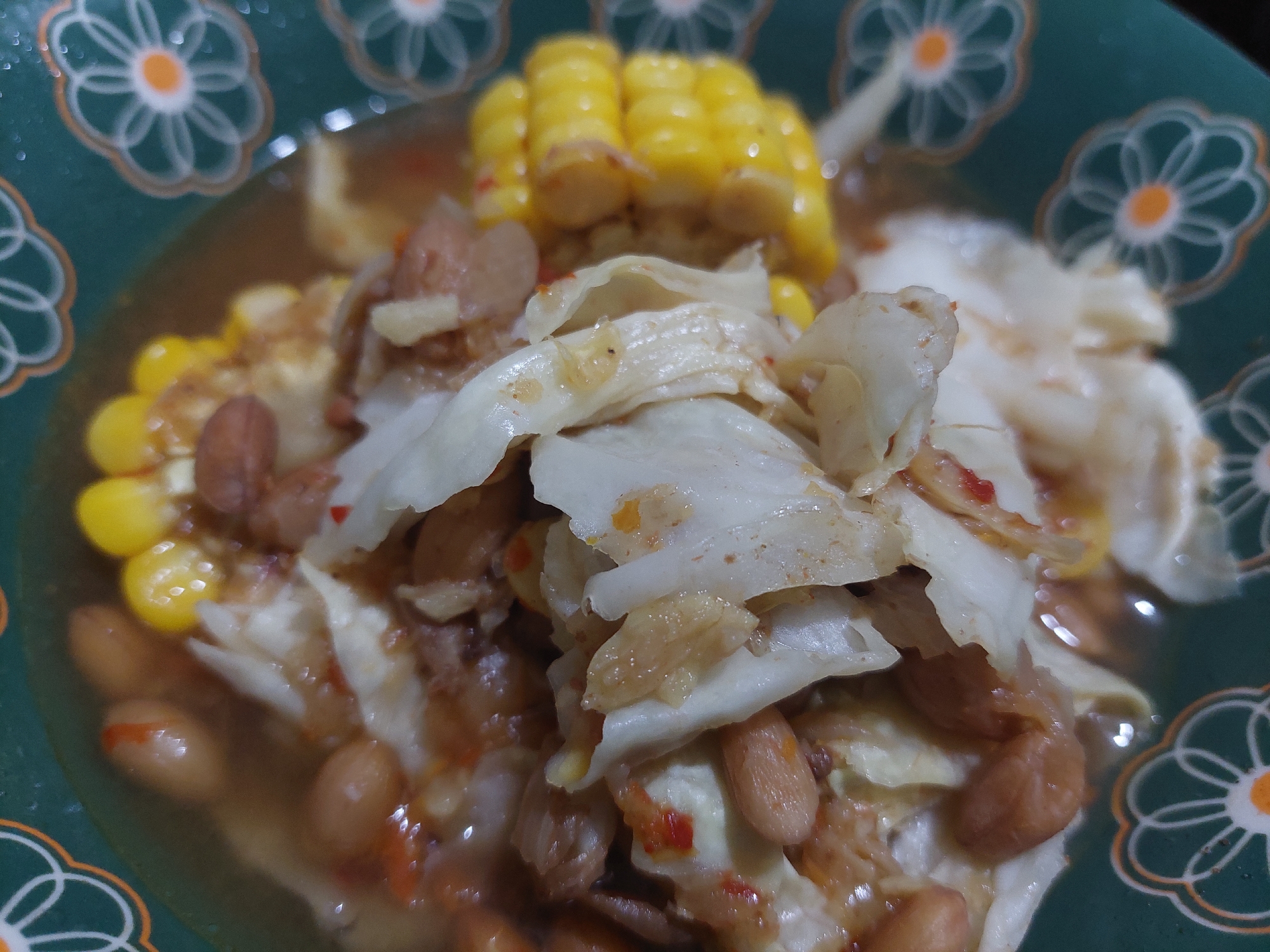 A close-up, overhead shot showcases a bowl of food with a green, floral-patterned rim. The food appears to be a stew or soup containing pieces of corn on the cob, roughly chopped white cabbage, and several peanuts in a brownish broth. The lighting is dim, and the focus is sharp on the food, with the background slightly blurred. The specific location in the city of Jelutung, Indonesia, is not discernible from the image.