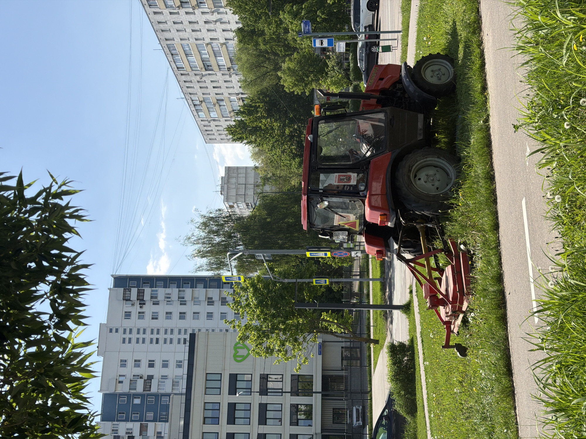 A red tractor with a connected mowing attachment is parked on the grass next to a sidewalk and asphalt road in what appears to be a city. Tall apartment buildings are visible in the background, and power lines stretch across the sky. The tractor appears to be a Belarus tractor, and the location is Moscow, Russia. Street signs are also visible near the tractor.