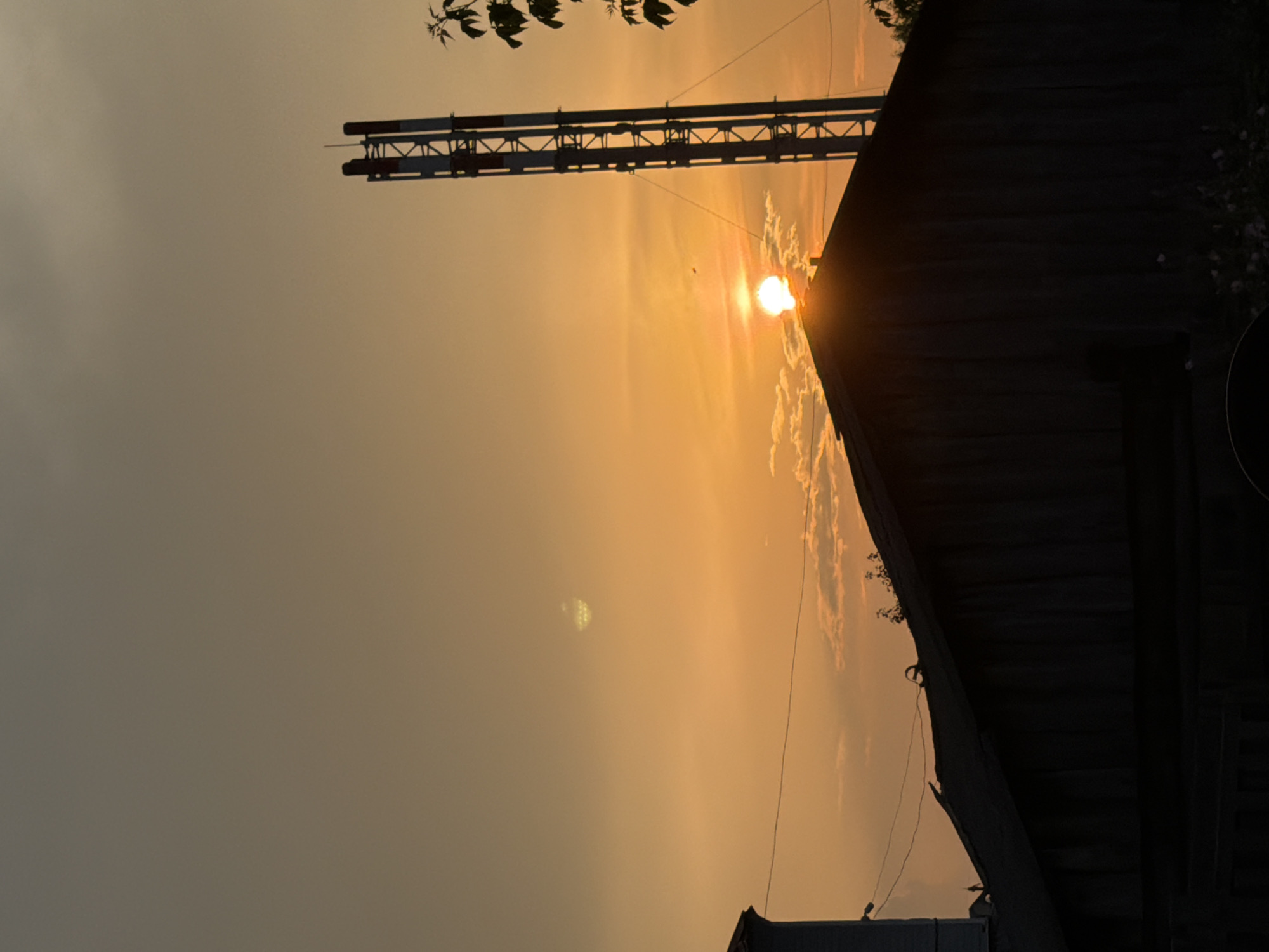 A silhouetted view of buildings and a tall industrial structure against a dusky sky in Vikulovo, Russia, as the sun sets. The sun, a bright orb, is partially obscured by the peak of a dark, wooden roof in the foreground. To the right, a tall, metallic tower with red and white markings rises vertically. The sky is a warm gradient of orange and yellow, with scattered clouds. A dark branch with leaves is visible on the far right. The image captures a tranquil yet slightly industrial atmosphere, characteristic of a small town at dusk.