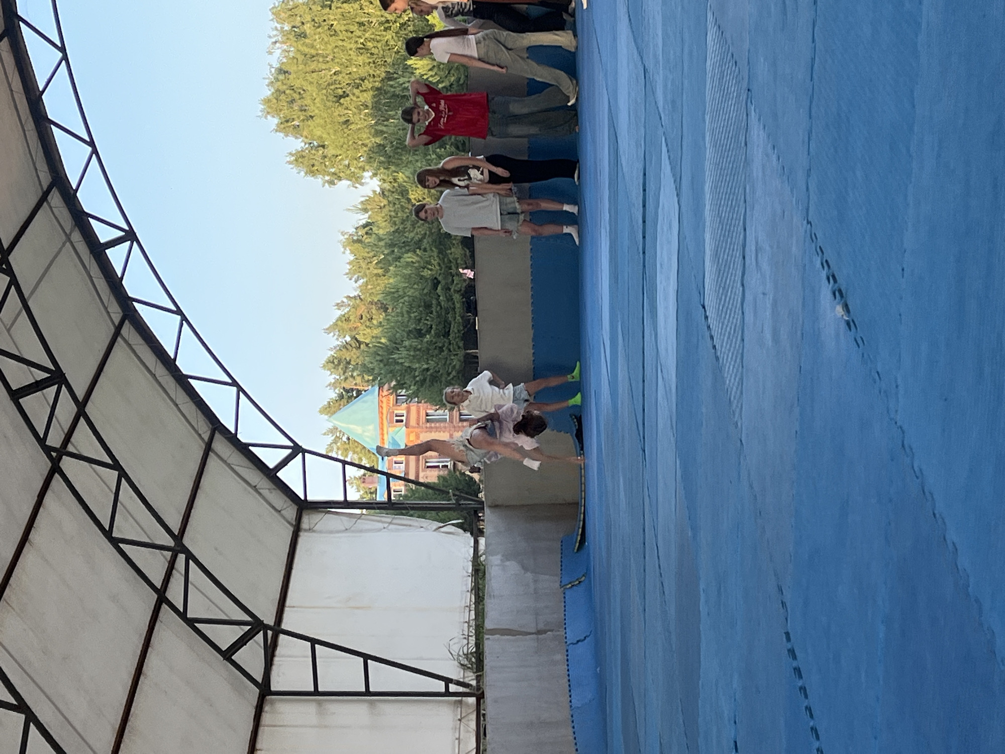 In an outdoor setting in Krasnoturansk, Russia, a group of young people are engaged in what appears to be a gymnastics or acrobatic training session. The scene is set on a bright, clear day, with a blue sky visible above. The ground is covered with large, interlocking blue mats, typical of a training facility.
A structure with a white canopy and a metal frame is visible to the left, suggesting an outdoor training area or a covered section of a larger facility. In the background, lush green trees and a building with distinctive green-tiled roofs and brickwork provide context to the location.
Several individuals are captured in various poses. One person is performing an acrobatic move, with their body in a dynamic, arched position, legs extended. Another individual, wearing a white shirt and shorts, appears to be assisting or spotting them. Further back, a line of people are standing, some in casual athletic wear, watching the activity. Their stances suggest they are either waiting for their turn, observing, or possibly participating in warm-up exercises. The overall mood seems to be one of active training and camaraderie among the participants. The time of day is likely afternoon, given the bright lighting and the clear sky.