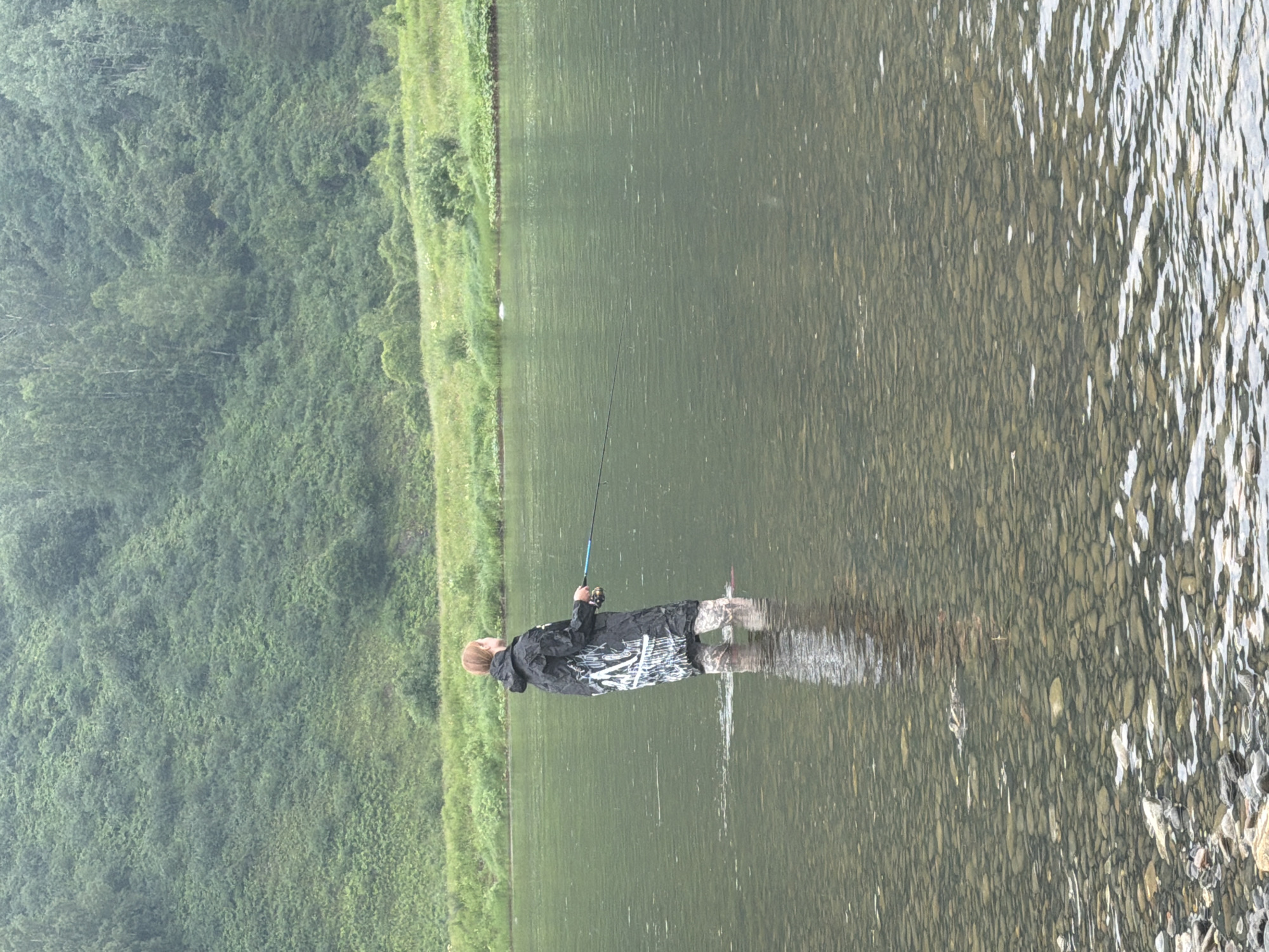 A person stands in the shallow water of a river, holding a fishing rod. They are wearing waders and a dark, hooded jacket with white markings. The riverbed is visible, covered in stones. In the background, a lush, green hillside rises steeply, covered in trees and vegetation. The sky appears overcast. The location is the city of Ust-Kabirza, Russia.