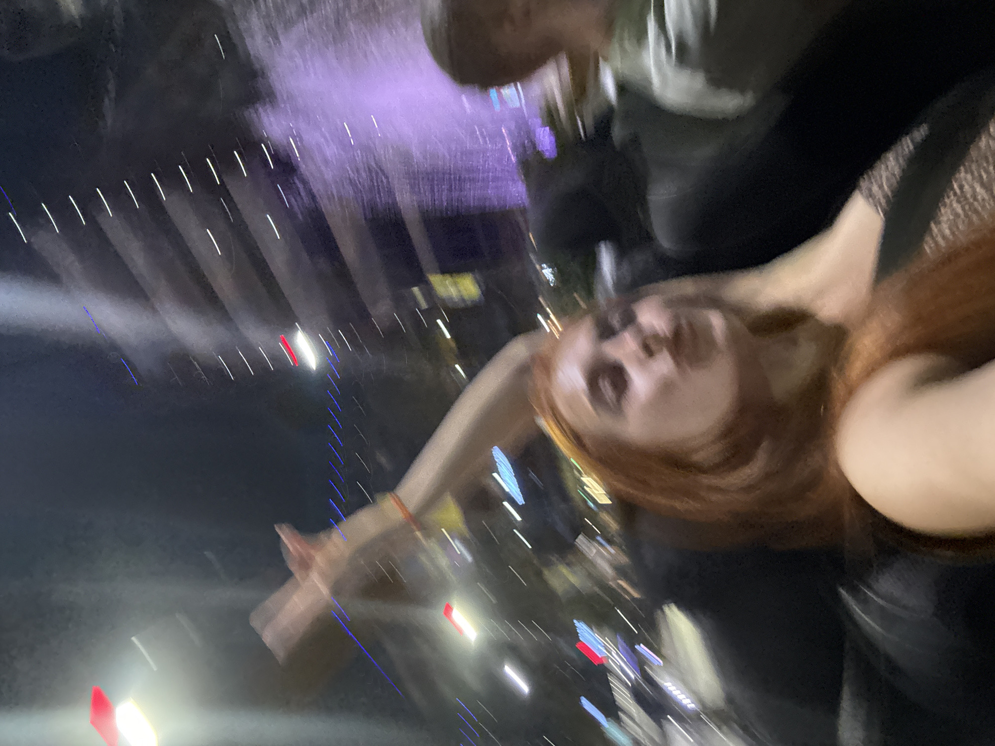 This is a blurry, low-light, wide-angle selfie taken at night. The primary subject is a young woman with long, reddish-brown hair, who is positioned in the lower right of the frame and looking towards the camera. Her face is partially visible, appearing to smile or laugh. She is wearing a dark, possibly black, outfit with a textured pattern on the shoulder or neckline, resembling fishnet or lace.
Behind and to the left of the woman, another person is visible, though their features are heavily blurred due to the low light and motion. This person appears to be wearing a light-colored shirt or jacket.
The background is a chaotic blur of lights and abstract shapes. Streaks of white and purple lights dominate the upper portion of the image, suggesting artificial lighting, possibly from a fountain, stage, or light show. There are also scattered red, blue, and yellow lights, indicative of a bustling urban or event setting at night. The overall impression is one of movement and a lively atmosphere.
Given the location context of Budva, Montenegro, and the nighttime setting with colorful lights, this photo likely captures a moment at an outdoor event, festival, or possibly a lively street scene in the city. The blur suggests the photo was taken quickly or during a period of motion, adding to the sense of spontaneity and fun. No discernible text is visible in the image.