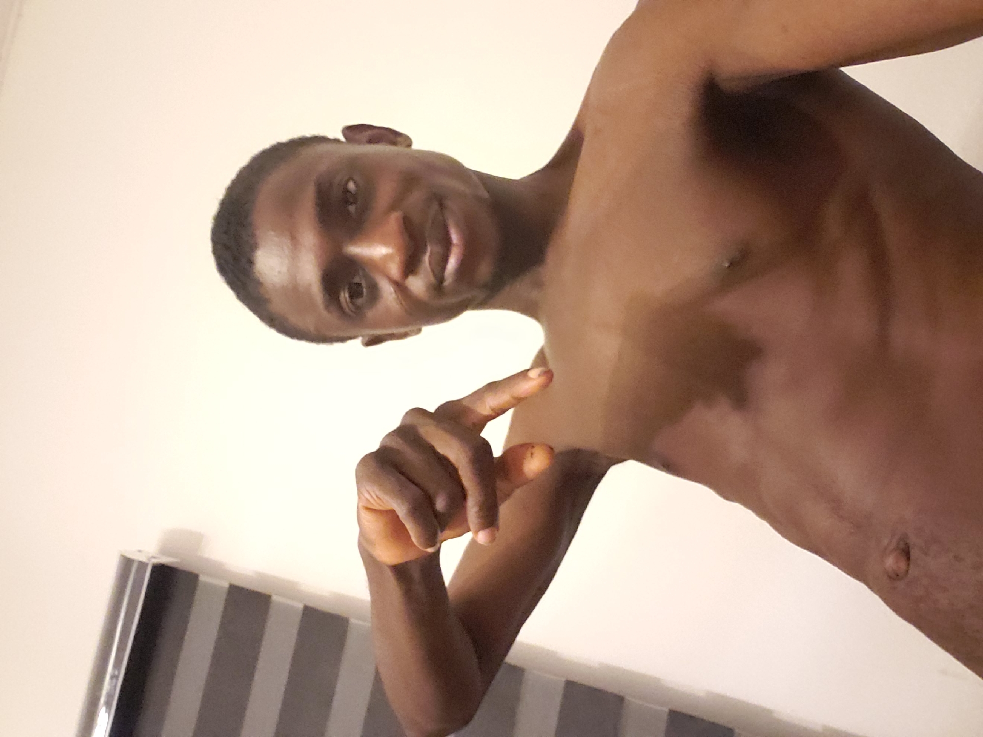 A medium shot depicts a shirtless man with dark skin, facing the camera at an angle, with his right hand raised, index finger extended and thumb bent. He has short, dark hair. A small portion of a window with horizontal blinds is visible in the lower left corner of the frame. The background consists of a plain, light-colored wall. The location is purportedly Adonkolo, Nigeria, although this cannot be verified from the image.