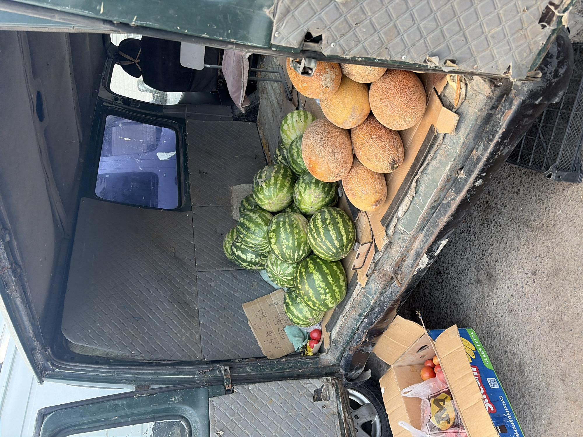 The image depicts the interior and exterior of an open vehicle, possibly a truck or van, filled with various fruits. In the back of the vehicle, a collection of cantaloupe melons with textured orange skin are piled up. Behind the melons, several watermelons with dark green stripes are stacked. Some of the watermelons are partially obscured by other fruits or the vehicle's interior. A small, worn cardboard sign with what appears to be handwritten text in a non-Latin script rests among the watermelons. Two red fruits, possibly apples or tomatoes, are also visible near the bottom of the watermelon pile.
To the right of the vehicle, a cardboard box lies open on the ground. The box is labeled with text, including "PREMIUM BANANAS" and possibly other branding information in a blue and yellow color scheme, suggesting it may have previously contained bananas or is intended for fruit transport. Inside the box, several red fruits, likely tomatoes or apples, are visible along with a plastic bag containing items that are difficult to discern.
The vehicle itself appears weathered and utilitarian, with a dark green exterior and a worn interior featuring textured gray paneling. The open door of the vehicle reveals a glimpse of the driver's side, including a seat and a window with a blue tint. The setting appears to be outdoors, on a paved or asphalt surface, suggesting a market or roadside stall. The lighting indicates it is daytime. The overall scene suggests a commercial activity, likely the sale or transport of fresh produce. There are no people visible in the image.