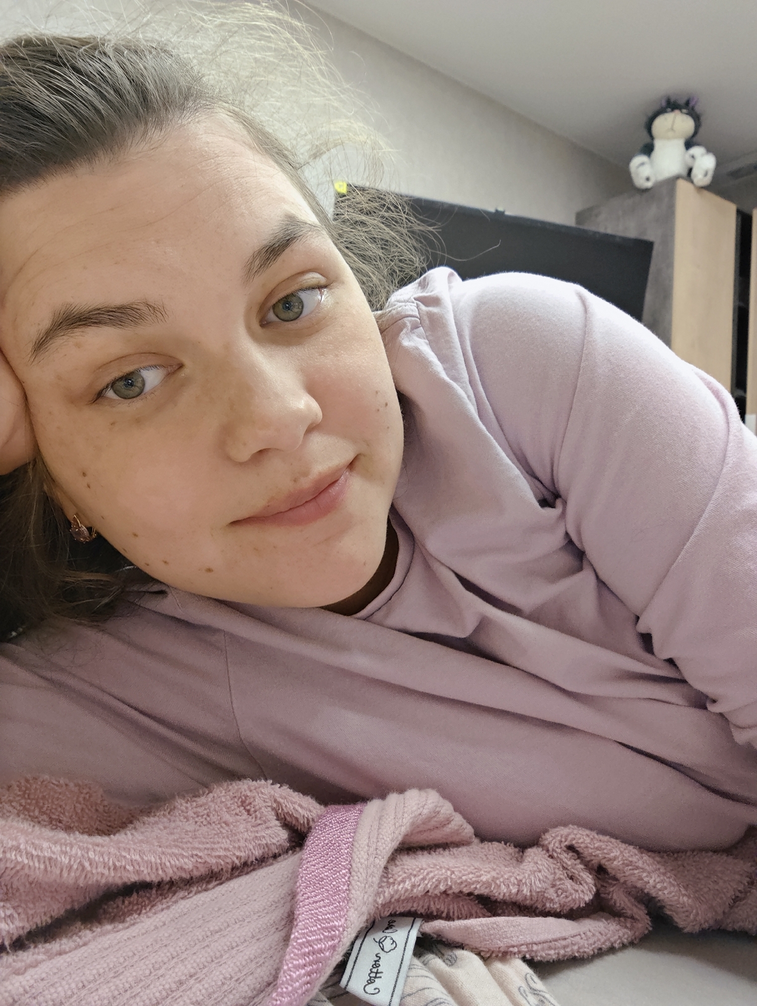 A young woman is captured in a close-up, laid-back portrait, likely taken indoors in Chaikovsky, Russia. She is lying on her side or stomach, resting her head gently on her right arm, which is partly visible. Her hair, a light brown shade, appears slightly dishevelled, suggesting a moment of relaxation or just waking up. She has fair skin, noticeable freckles or moles scattered across her face, and striking green or hazel eyes that meet the camera's gaze with a soft, slightly smiling expression. Her lips are naturally colored, hinting at minimal or no makeup.
She is wearing a comfortable-looking light lavender or pale purple long-sleeved top. In the foreground, below her, are layers of soft textiles: a textured, plush pink towel or blanket, and another lighter, possibly striped, fabric. A small white tag on one of the pink fabrics is partially visible, showing the word "cotton" and what appears to be a stylized logo.
The background is softly blurred but reveals an indoor setting, possibly a bedroom or living room. On a shelf or piece of furniture in the upper right corner, a small, black and white plush toy, resembling a cat or a cartoon character, sits upright. The lighting is soft and even, typical of indoor ambient light, contributing to the calm and intimate atmosphere of the image. The overall mood conveyed is one of ease, quiet contemplation, and contentment.