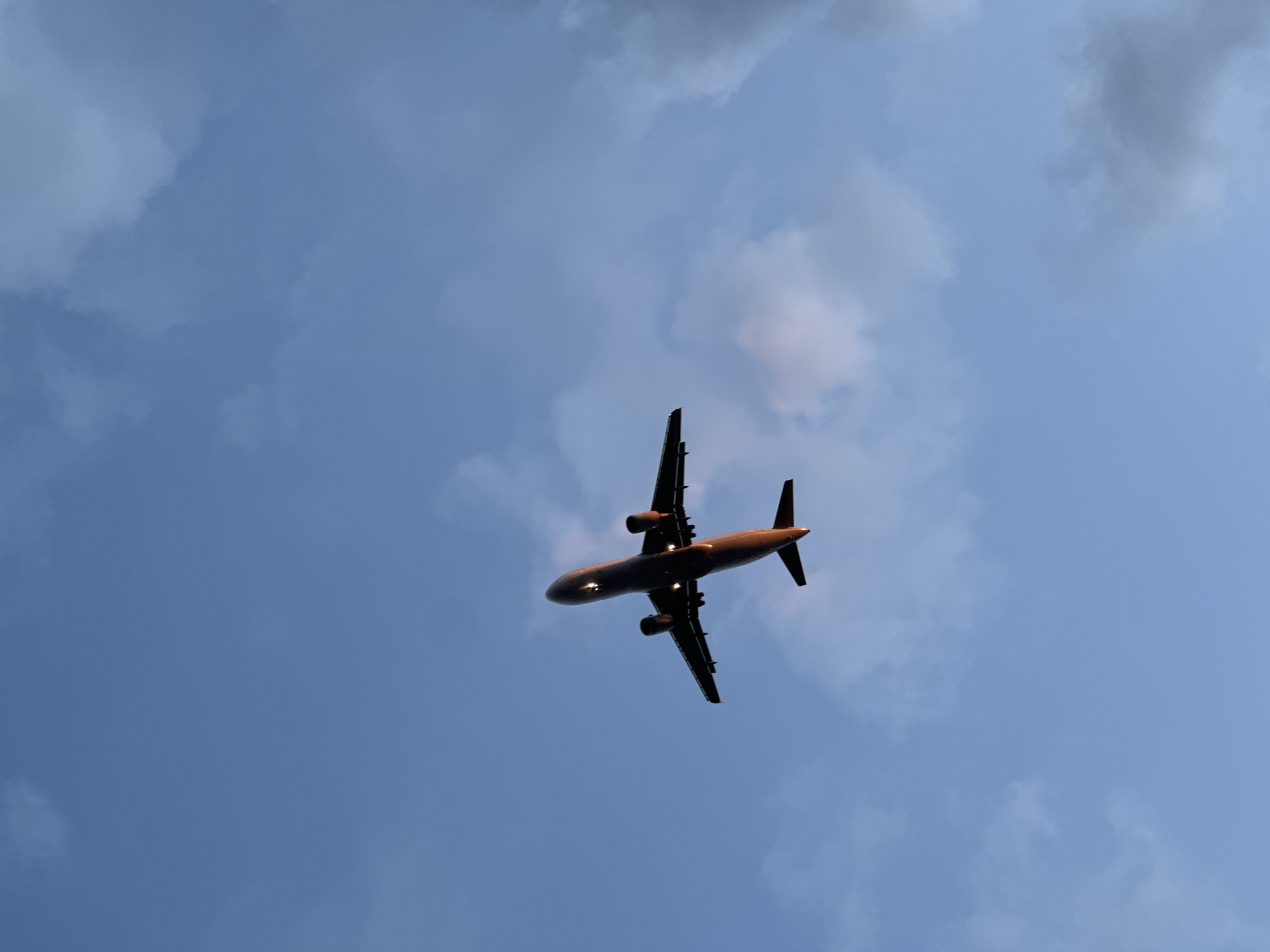 A twin-engine passenger jet aircraft is prominently featured against a blue sky, captured from a low-angle, upward perspective. The aircraft is dark, largely silhouetted, indicating the sun is behind or to its side, likely during sunrise or sunset hours. Warm, golden-orange light illuminates the underside of its fuselage and engine nacelles, highlighting its contours. Bright white landing lights are visible on the wing leading edges, and several illuminated cabin windows suggest interior lighting is active. The wings show extended flaps, characteristic of either takeoff or landing phases of flight, with the aircraft appearing to be in an ascending trajectory. The sky transitions from a lighter, clearer blue in the upper left to a deeper blue with scattered, varying densities of clouds towards the right and bottom of the frame. These clouds range from thin and wispy to slightly denser, grey formations, particularly on the right. The scene depicts the aircraft in flight over the city of Sofia, Bulgaria, likely in the vicinity of Sofia Airport (LBSF), given its apparent low altitude and flight configuration. No people or ground objects are visible, emphasizing the aircraft as the sole subject against the expansive sky.