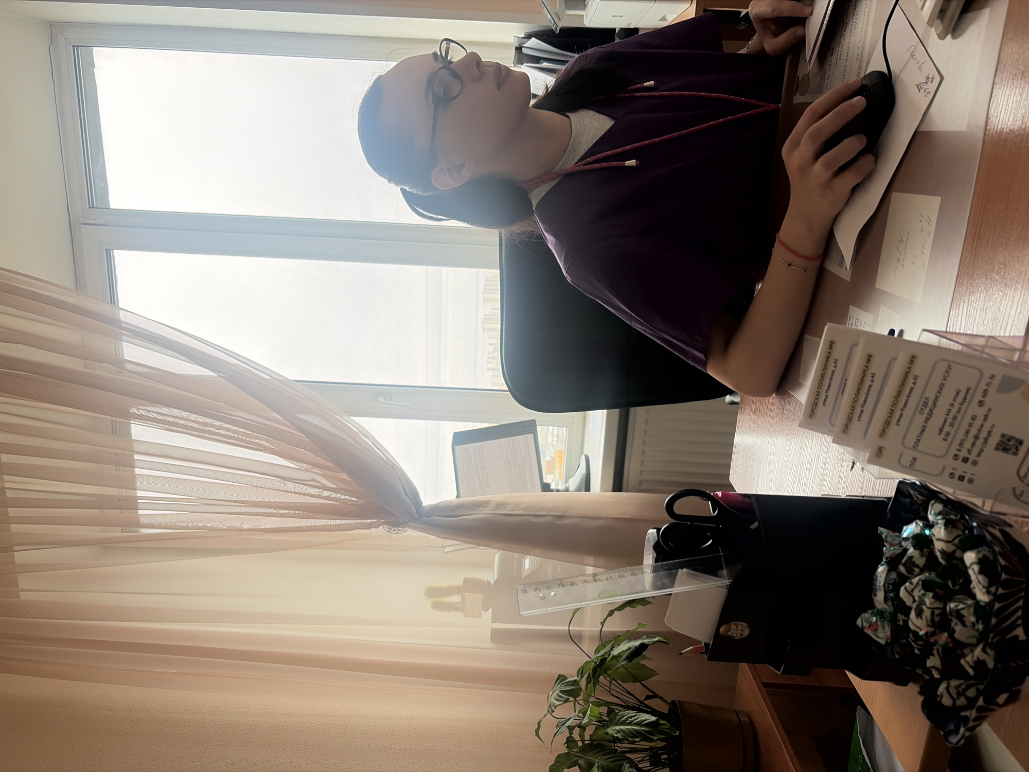 A young woman, likely in her late teens or early twenties, is seated at a wooden desk in an office setting. She has dark hair styled back, wears glasses, and is dressed in a purple top over a light undershirt, with a red cord featuring golden tips around her neck. A red string bracelet adorns her left wrist. Her right hand rests on a black computer mouse, suggesting she is actively working on a computer or reviewing documents on the desk. She appears focused on her task.
The desk is cluttered with various office supplies, including stacks of papers, a black desk organizer holding scissors and pens, a clear ruler, and a small potted plant with green leaves. A container filled with individually wrapped candies is also visible on the desk.
Behind the woman is a large window with white frames, letting in abundant, diffused natural light, indicating it is daytime with an overcast sky. Light brown, translucent sheer curtains are drawn, partially obscuring the view outside but still allowing light to filter through. Through the curtains, part of a computer monitor can be seen. In the background, behind the woman, shelves hold what appears to be a printer or scanner.
Several documents on the desk feature visible Russian text, identifying them with "ГОРОДСКАЯ ПОЛИКЛИНИКА №8" (City Polyclinic No. 8) located at "улица Новосёлов, д.45" (Novoselov Street, building 45). One document specifies the "ОТДЕЛ ПЛАТНЫХ МЕДИЦИНСКИХ УСЛУГ" (Department of Paid Medical Services) in "кабинет 414 (4 этаж)" (Cabinet 414, 4th floor), with operating hours listed as "8:00 - 20:00 (по будням)" (8:00 - 20:00 on weekdays) and contact phone numbers provided. These details indicate the scene is likely an administrative area of a medical facility, specifically a polyclinic, in Saint Petersburg, Russia.