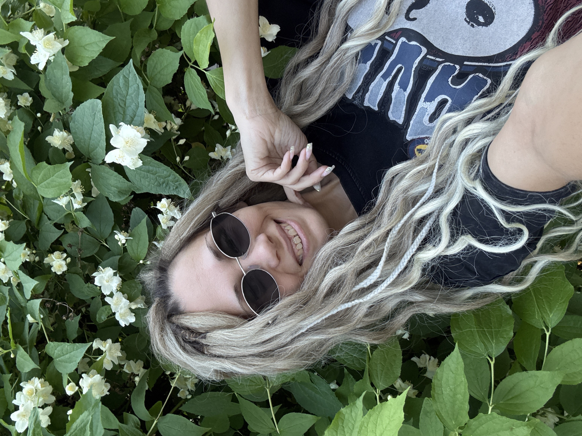 An outdoor self-portrait features a smiling female individual positioned against a dense backdrop of green foliage interspersed with numerous white, five-petaled flowers, indicative of a flowering shrub, in the city of Rusanovo, Russia. The subject's head is slightly tilted to her left, and she is looking directly at the camera. She wears dark-lensed, oval-shaped sunglasses with thin silver frames. Her long, wavy hair is light blonde, with several sections styled as dreadlocks or thick braids, and includes distinct thin white braids. Her smile reveals visible teeth, with the upper front teeth exhibiting a slightly darker or discolored appearance. Her right hand is raised, with fingers adorned with elongated, light-colored or glittery manicured nails, gently resting near her chin. She is wearing a dark, short-sleeved t-shirt that features a graphic design comprising large, stylized blue and white letters (partially visible, possibly "CAL") alongside a circular motif that appears to be a cartoon character or symbolic figure, rendered in blue, white, and reddish-brown tones. The scene is brightly lit by natural, diffused light, indicating daytime.