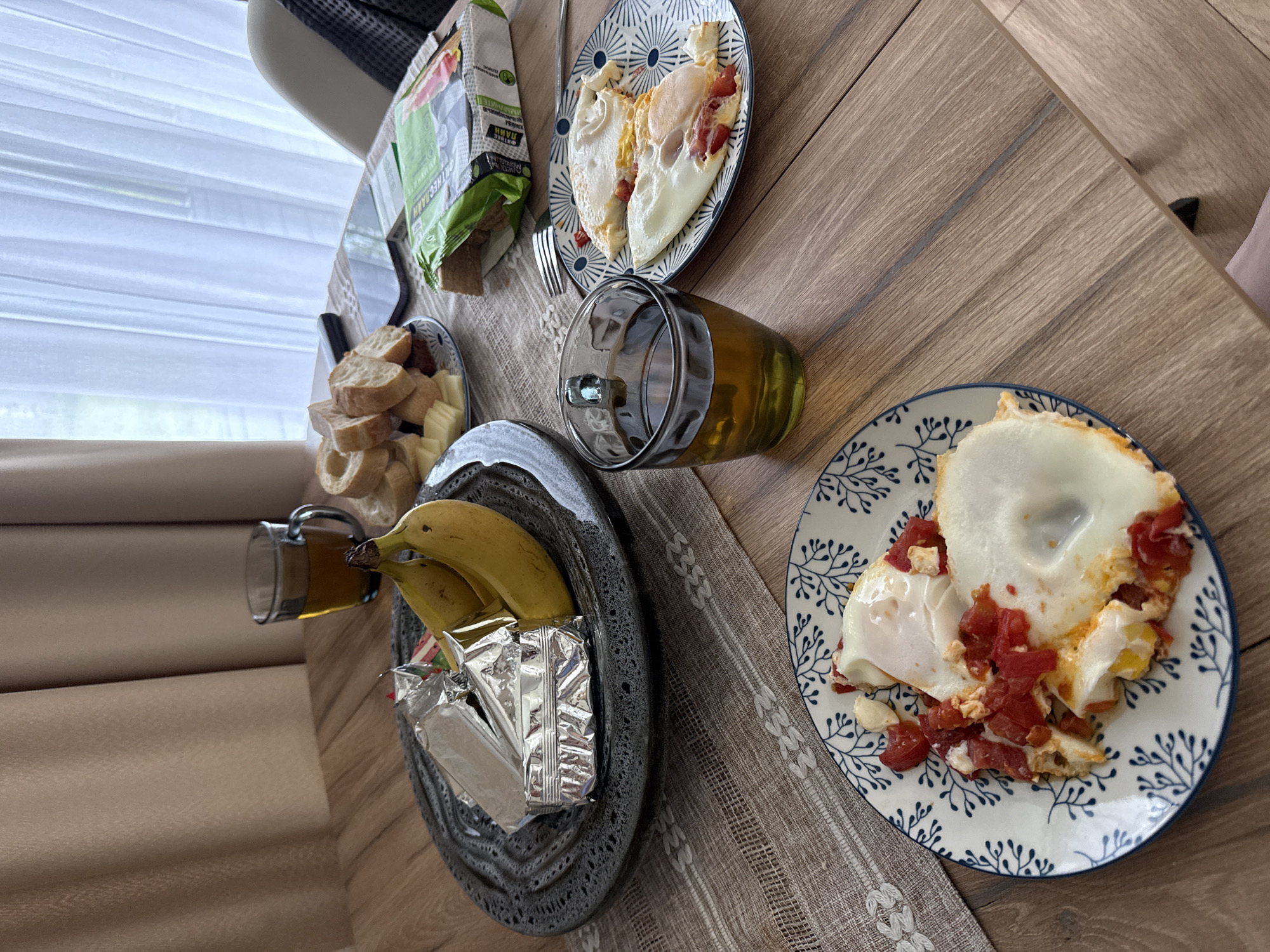 The image depicts a breakfast scene on a wooden table. A table runner with a geometric pattern is beneath the food. On the left side of the table, there is a dark gray ceramic plate with bananas and wrapped cheese. Next to it, a glass mug filled with liquid. A plate with bread slices and cheese cubes sits further up the table. On the right side are two plates of sunny-side-up eggs with tomatoes. Behind the left plate, there is a bag of chips. A fork is next to the plate with the chips. The location is Buzhaninovo, Russia.