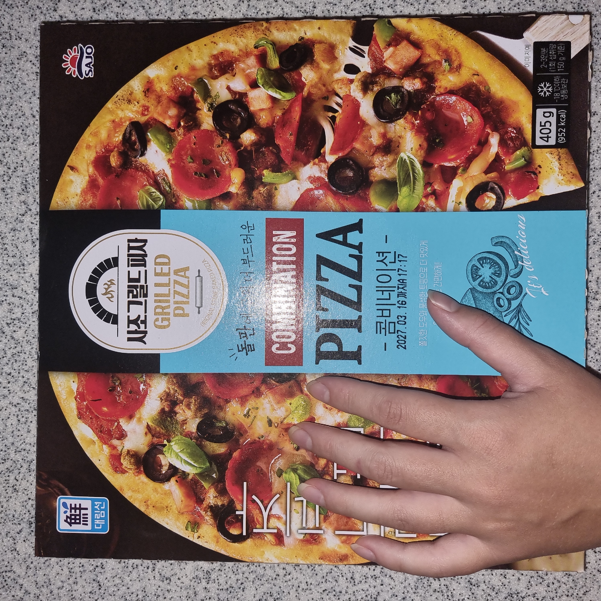 The image is a close-up, top-down view of a pizza box. The box features a large, appetizing image of a combination pizza with pepperoni, olives, green peppers, and basil. Text on the box is primarily in Korean, with "SAJO GRILLED PIZZA" prominently displayed. There are also English words like "COMBINATION PIZZA" and "ORIGINAL STONE BAKED PIZZA". A hand is partially obscuring the bottom left portion of the box. In the bottom right corner, there is nutritional information including weight (405g) and calorie count (952 kcal), along with storage instructions and serving size. The box is placed on a speckled, greyish-white surface, resembling a countertop. The city of Dunnae-myeon, South Korea is not visible or suggested in the image.