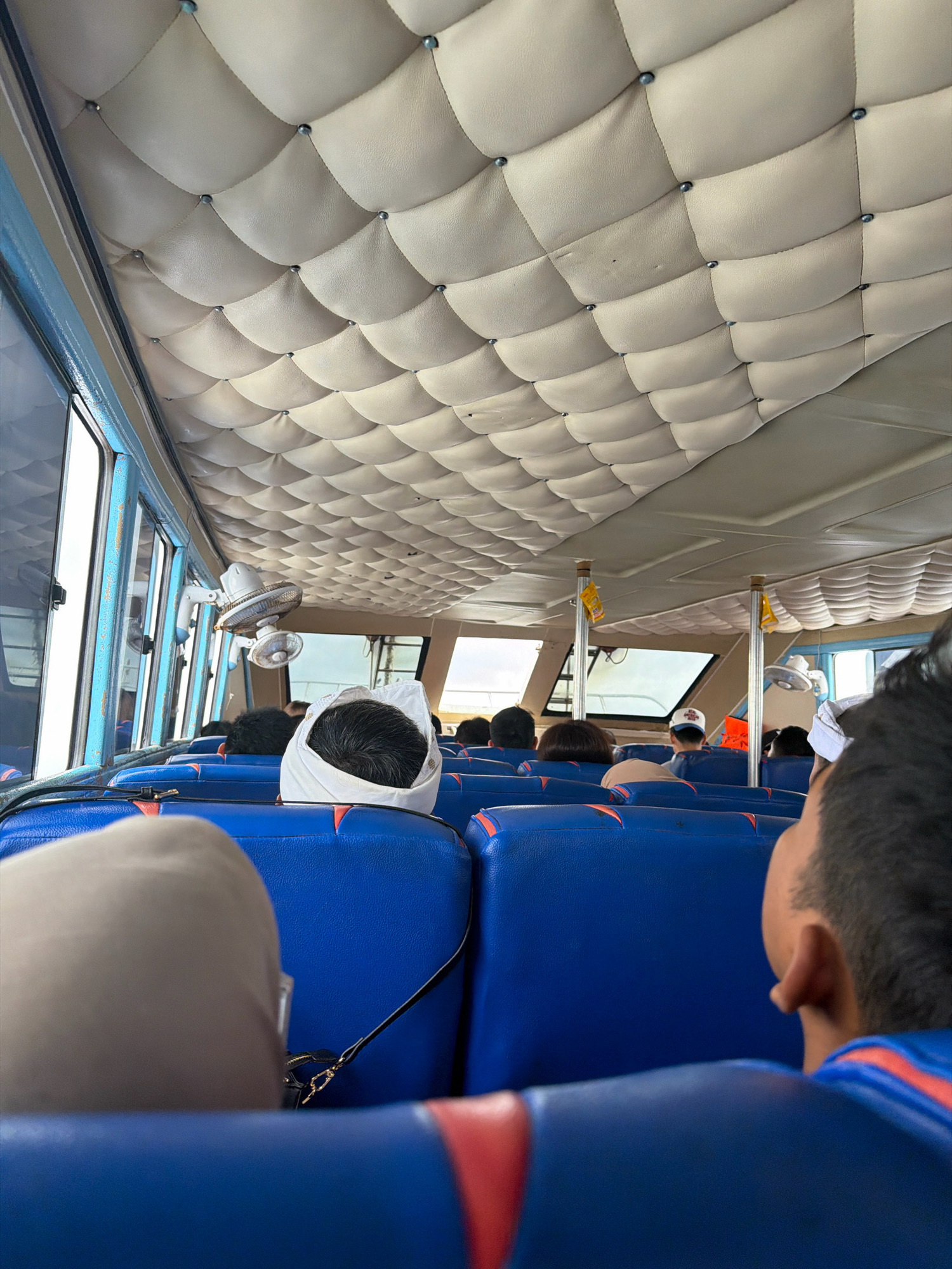 The image shows the interior of a passenger boat, likely in Unknown, Indonesia, judging by the presence of a ceiling fan and the general aesthetic of public transportation in the region. The ceiling is upholstered with a tufted beige material, creating a diamond pattern with silver studs at each intersection. Blue and red upholstered seats are visible, arranged in rows. Several passengers are seated, with their backs mostly to the camera. One person in the foreground to the left is partially visible, wearing a tan head covering. Further back, a person wearing a white head covering is prominent. In the mid-ground, two white electric fans are mounted on the wall. The windows along the side of the boat offer a glimpse of the outside, which appears to be bright and possibly overcast. The overall impression is one of travel or commuting within a local Indonesian setting.