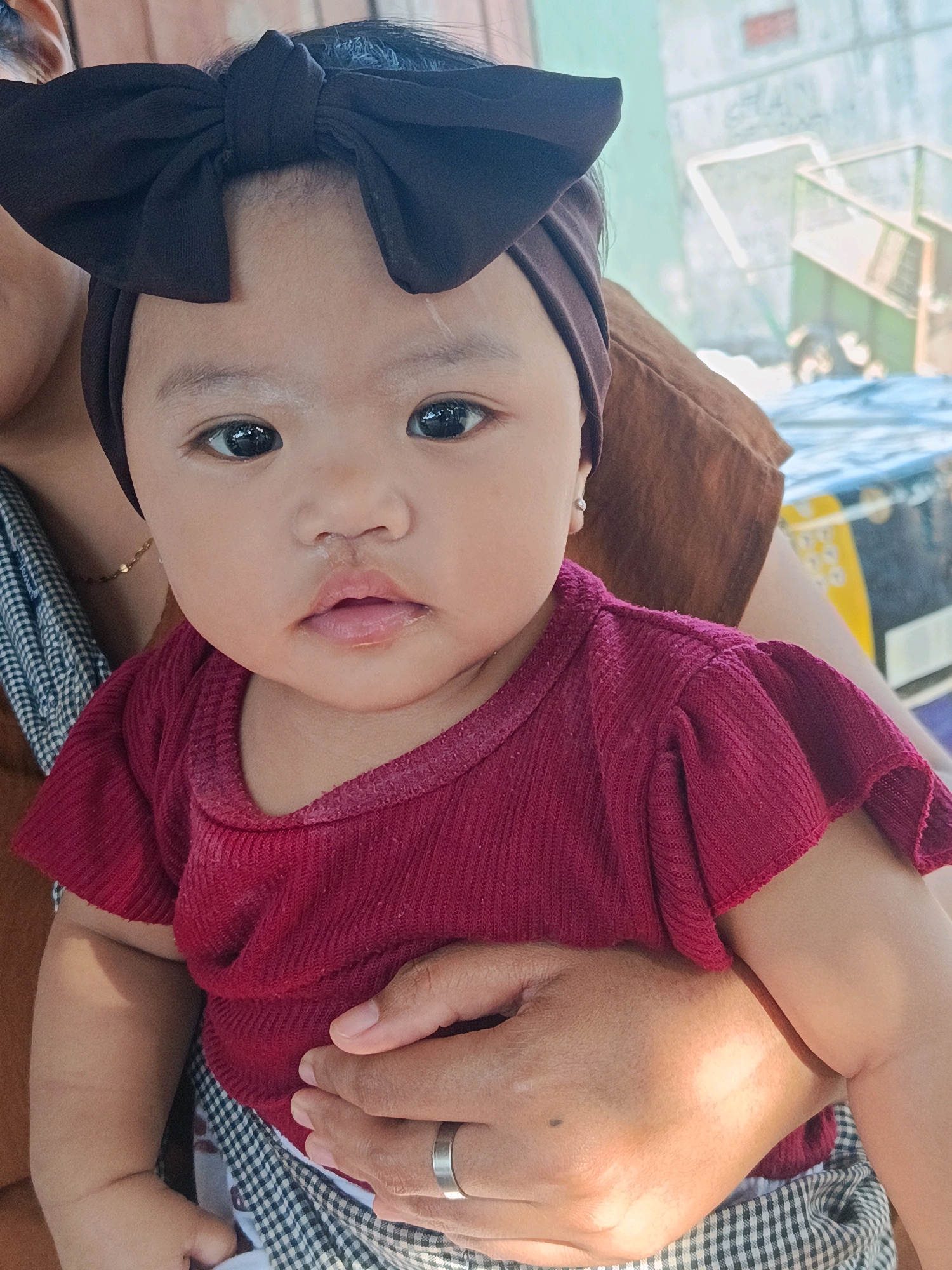 A baby, wearing a dark brown bow headband and a maroon ruffled-sleeve shirt, is held by an adult's arms. The baby's eyes are open and looking forward, and their lips are slightly parted. The adult's hands are visible, one resting on the baby's chest and the other supporting their legs. The background appears to be an outdoor setting with a blue tarp and some structures, possibly in the city of Tempeh, Indonesia.