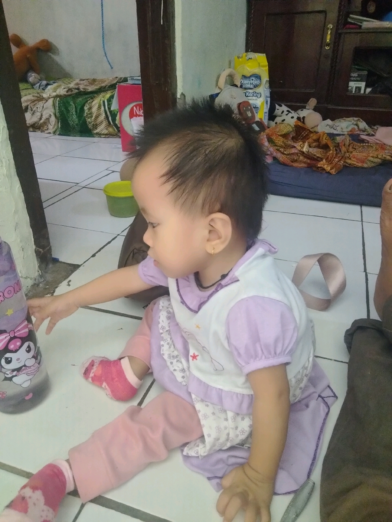 A toddler is sitting on a tiled floor in what appears to be a room in a residential setting in Suka Sari, Indonesia. The toddler is wearing a light purple and white dress with a pink underskirt and pink socks. One of the toddler's legs is bandaged with pink and white material. The toddler's left hand is reaching towards a purple bottle with a cartoon character on it. In the background, there are several items including a red and white box with the word "Nabi" on it, a yellow bag of diapers, and some stuffed animals. The floor is tiled with a white and gray pattern. The lighting suggests it is daytime.