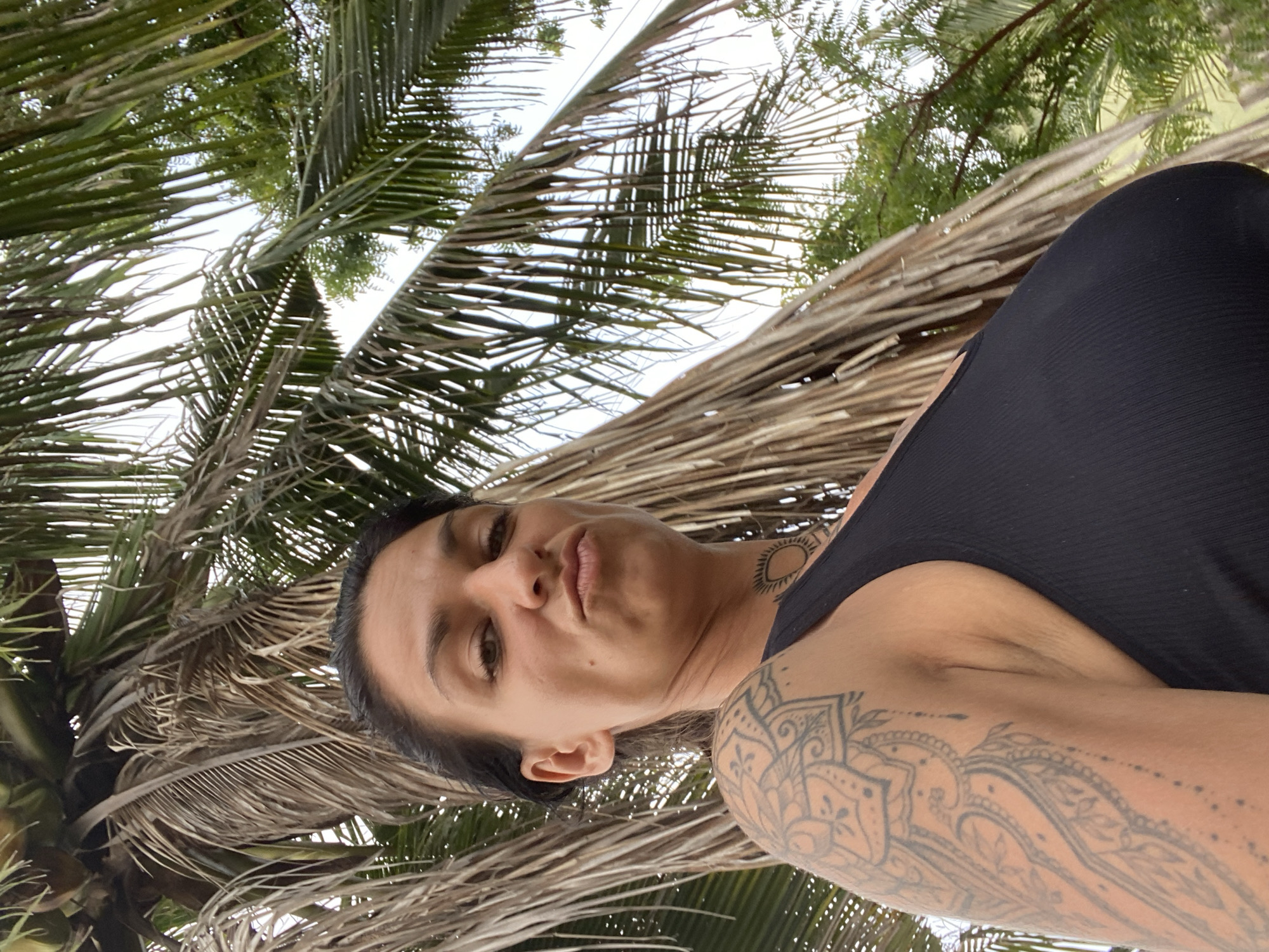 In Mũi Né, Vietnam, a woman with dark hair and a tattoo on her left arm and neck is pictured under a palm tree. She is looking at the camera with a playful expression, her lips pursed. The palm fronds are dry and brown, with green foliage visible in the background. She is wearing a black ribbed tank top. The sky is overcast.