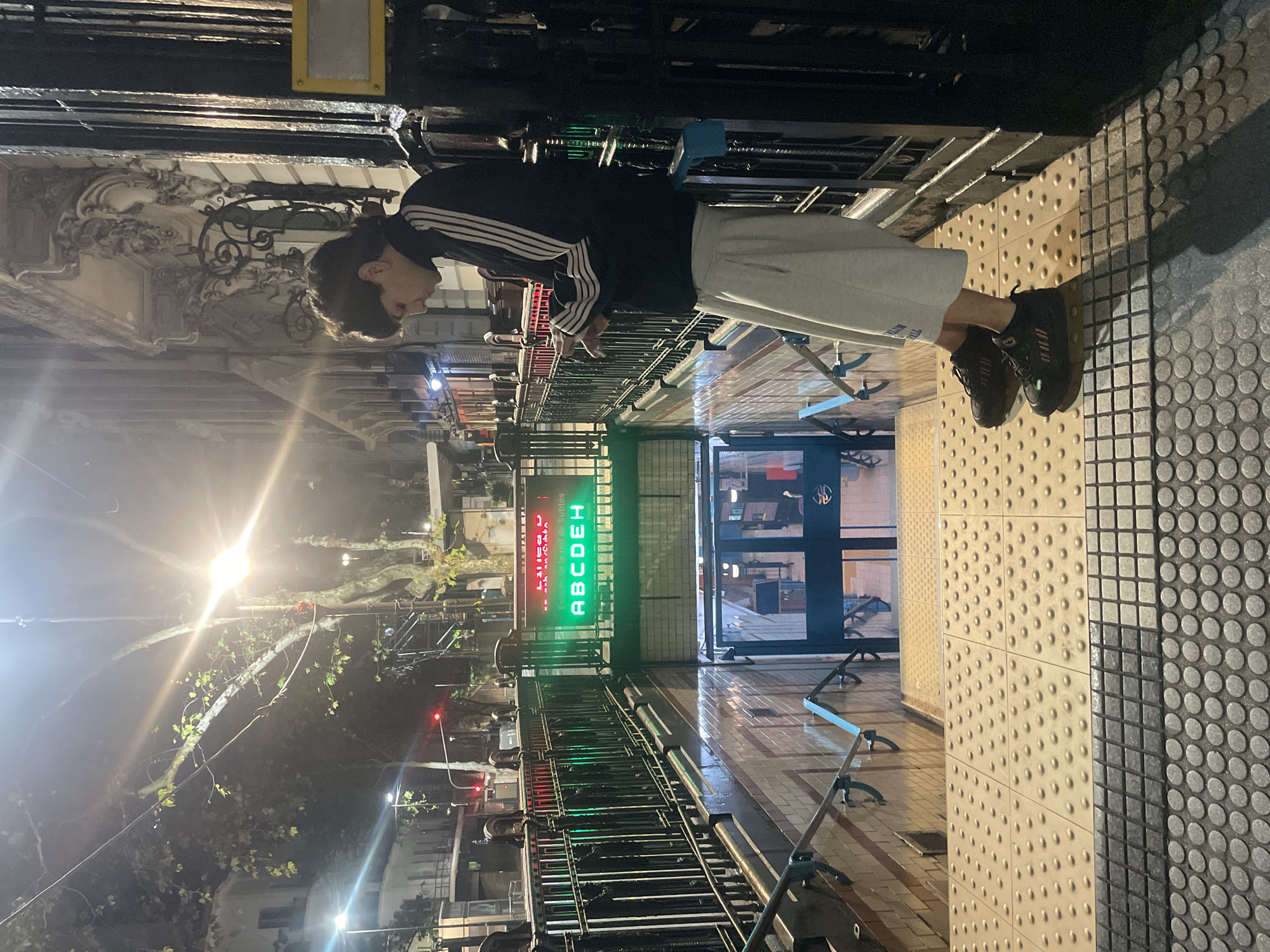 The image captures a nighttime urban scene, focusing on a young person standing near the entrance of a subway (Subte) station. The person, appearing to be male, is dressed in a dark track jacket with white stripes (Adidas-style), light-colored shorts, and dark athletic shoes. He has short, dark hair and is looking downwards, possibly at a phone or the ground.
The background reveals the entrance to the "Subte," featuring metal turnstiles. Above these are glowing LED signs. One prominent sign displays "SUBTE" in red text and "ABCDEH" in bright green, indicating the operational subway lines. Another red sign, partially visible, reads "Estacion: Plaza de Mayo" and "COMBINACION," identifying the station and a transfer option. Beyond the turnstiles, an open entrance leads to a tiled interior, suggesting the station platform.
The ground around the entrance features tactile paving: light-colored tiles with raised circular dots for visual impairment, contrasting with darker, square-patterned tiles. In the broader background, a bright streetlamp on the left illuminates tree branches with sparse green leaves against the dark night sky, causing a strong lens flare. Across the street, ornate architectural details of older buildings are visible, adding to the urban ambiance. The scene is clearly set at night, with artificial lighting providing the main illumination. The "Subte" and "Plaza de Mayo" references firmly place the location in Buenos Aires, Argentina.