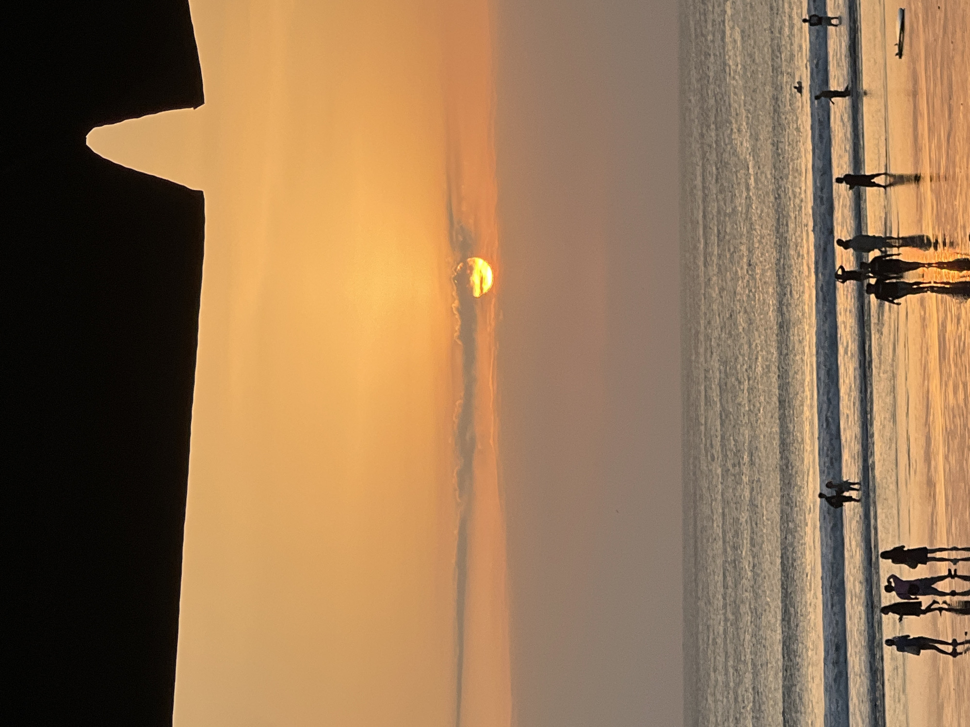 A sunset over the ocean in Kuta, Indonesia, is captured from a low angle, with a dark, silhouetted awning in the foreground. The sky is a vibrant orange and yellow, with the sun partially obscured by a thin layer of clouds. Several people are walking along the beach and wading in the water. Some are silhouetted against the bright sky, while others are more visible on the wet sand, their reflections mirroring the sunset. The ocean has gentle waves, and a surfboard is visible lying on the sand. The overall mood is serene and peaceful, with the warm colors of the sunset creating a beautiful and tranquil atmosphere.