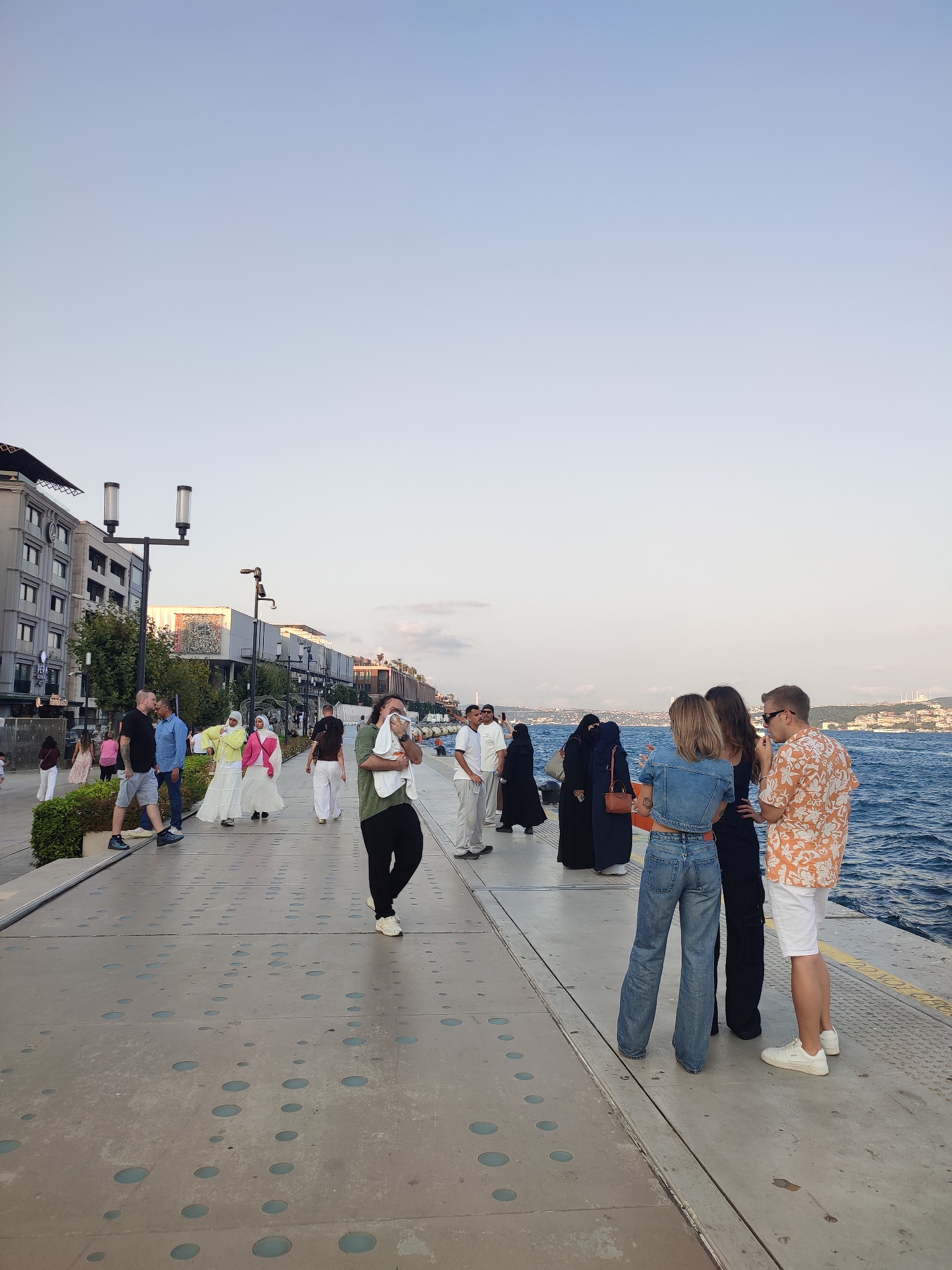 The image depicts a waterfront promenade in the Beyoğlu district of Istanbul, Turkey, with the Bosphorus Strait visible on the right. A group of people are strolling along the paved walkway, which features decorative circular indentations.
In the foreground, on the right side of the image, three individuals are standing and conversing. One man, wearing an orange and white patterned short-sleeved shirt and white shorts with white sneakers, is facing left. To his left, a woman in a denim jacket and wide-leg jeans is facing away from the camera, engaged in conversation with another woman whose back is also to the camera.
Further down the promenade, several other people are visible. Two individuals in black Islamic attire, including a niqab, are standing, and a woman in a yellow hijab and a white dress is walking. A man in a green t-shirt and black pants is in the mid-ground, holding a white cloth to his face. On the left side of the walkway, near a building with several windows and balconies, a man in a black t-shirt and grey shorts walks with another man in a blue shirt. Other pedestrians are scattered throughout the scene.
The environment includes a modern building complex with shops and restaurants lining the left side of the promenade. Trees and greenery are also present. The water of the Bosphorus is choppy, and in the distance, a cityscape with structures and a bridge can be seen. The sky is clear and pale blue. The overall atmosphere suggests a public space being utilized by both locals and tourists.
