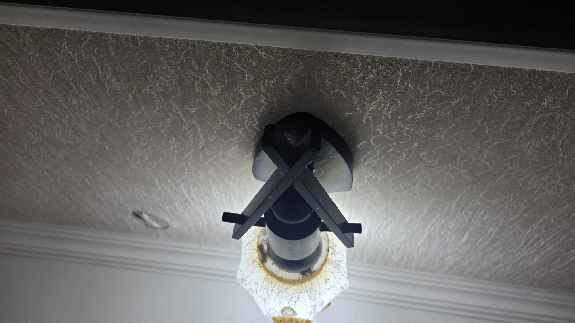 An ornate, dark brown wooden sconce holds a decorative light fixture with a cracked, white glass lampshade. The sconce is mounted on a textured, light beige wallpaper. The wallpaper has a subtle, irregular pattern resembling fine, organic lines. Above the sconce, a white, decorative ceiling molding is visible. The image is taken from a low-angle perspective, looking upwards towards the ceiling. In the upper right portion of the image, a light gray, oval-shaped mark is present on the wallpaper. The lower edge of the image shows a dark, shadowy area, possibly a floor or furniture. This scene is likely an interior, possibly a hallway or living area, in Chuy, Kyrgyzstan.