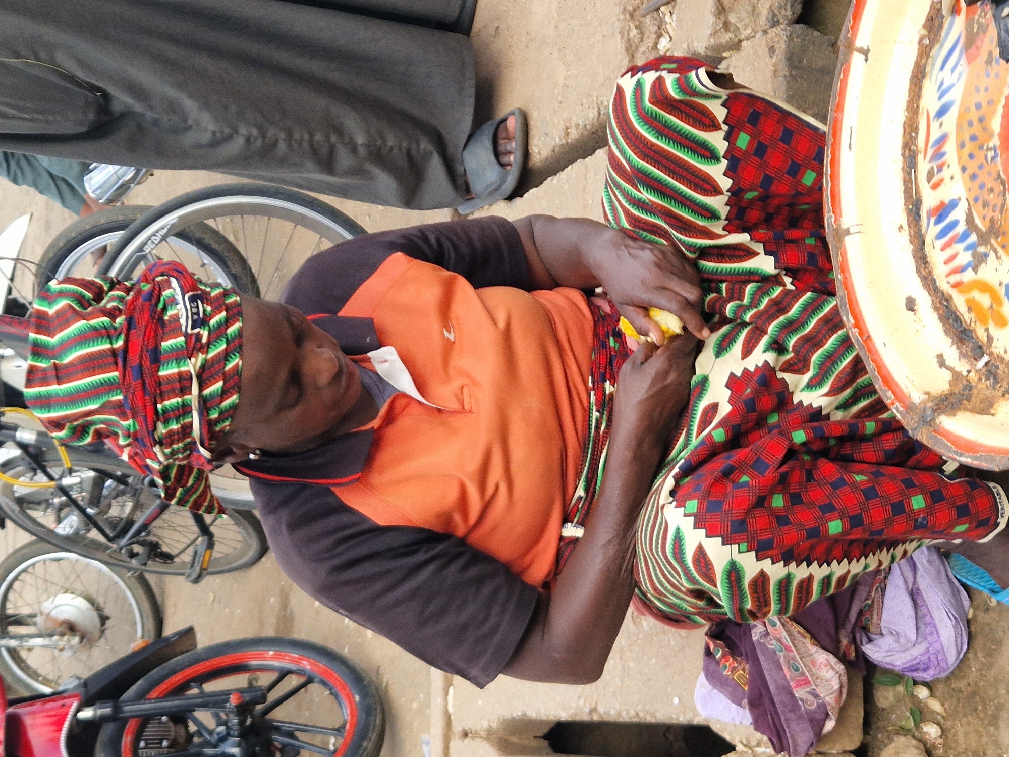 A woman sits on the ground with a brightly colored skirt and headwrap, peeling a piece of yellow fruit. She wears an orange shirt with black sleeves and has an earring in her visible ear. Next to her are stacks of fabric and a large metal bowl with a painted fish design. In the background, there are bicycles and a person in dark clothing wearing sandals. The ground appears to be dirt and concrete, with a drainage grate visible. The scene takes place outdoors, likely in a market area of Jos, Nigeria.