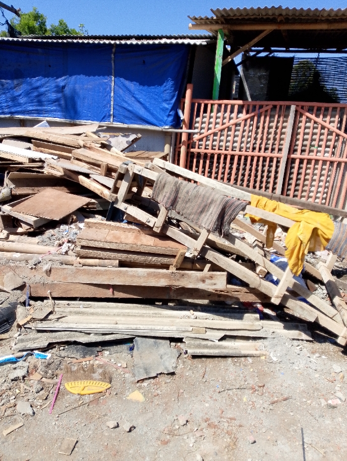 The image displays an outdoor scene characterized by a significant pile of assorted materials in the foreground and midground, set against a backdrop of improvised structures under a clear blue sky.
In the center and extending into the foreground, there is a large, haphazard pile primarily composed of wooden planks, beams, and what appear to be disassembled wooden frames or pallets. The wood varies in size and condition, suggesting it might be scrap material or salvaged lumber. At the base of this wooden pile, several pieces of corrugated metal sheets, possibly roofing or siding, are visible. Perched on top of the wooden debris are two distinct pieces of fabric: one is a dark, striped textile, possibly a rug or mat, draped over some wooden beams, and further to the right, a bright yellow piece of fabric, likely clothing or a towel, is also resting on the pile. The ground beneath the pile and extending into the foreground is a dusty, uneven surface of dirt and gravel, scattered with small pieces of debris including what looks like a yellow plastic object (possibly a toy or tool) and a pink plastic stick.
In the background, behind the pile of materials, several structures are visible. To the left, a building wall is partially covered by a large, bright blue tarp, which seems to be serving as a temporary or makeshift wall covering. Above this, a corrugated metal roof is visible. Further to the right in the background, another section of a structure features an orange-red colored fence or gate with a prominent grid-like pattern. This fence appears to be made of metal or painted wood. Above this section, another corrugated metal roof is present, supported by visible wooden or bamboo beams, one of which is painted green. A dark, possibly netted or fabric-like material is visible behind the orange fence. Above the structures, the sky is a clear, vibrant blue, indicating bright daylight conditions.
The overall scene suggests an environment where construction, renovation, or disuse is evident, possibly in an informal settlement or a backyard area where materials are stored or discarded. No people are visible, and no specific activity is taking place, presenting a static snapshot of the area.