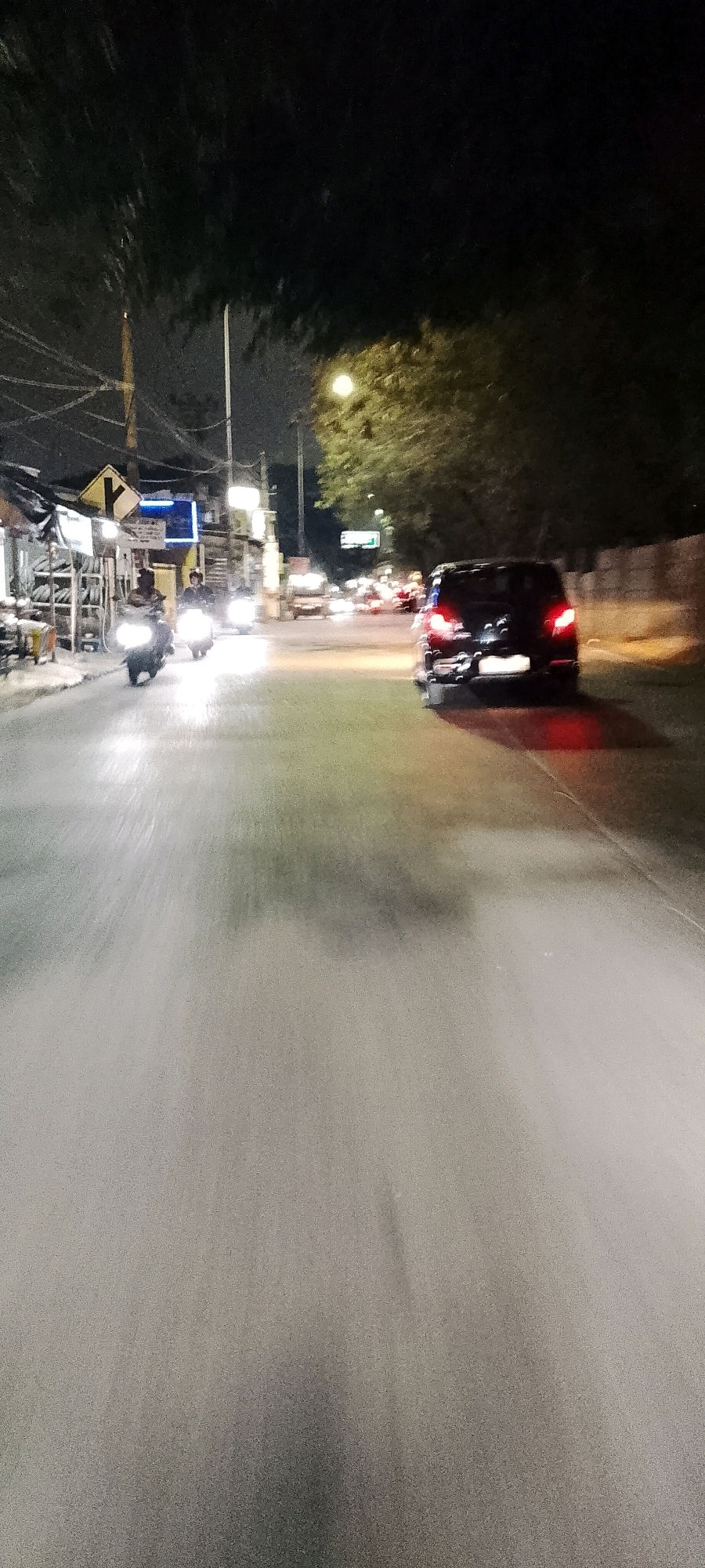 This is a night-time, motion-blurred photograph taken from a vehicle on a paved road in Depok, Indonesia. The scene depicts a moderately busy street with several vehicles, including motorcycles and cars. On the left side of the road, there are small shops or stalls with goods displayed. Traffic signals, streetlights, and overhead electrical wires are visible, suggesting an urban or suburban environment. The road surface is wet, reflecting the headlights of vehicles and streetlights, creating streaks of light. The trees lining the right side of the road are dark and leafy, with some illumination from the streetlights. The overall impression is a street scene at night with active traffic and commercial activity.