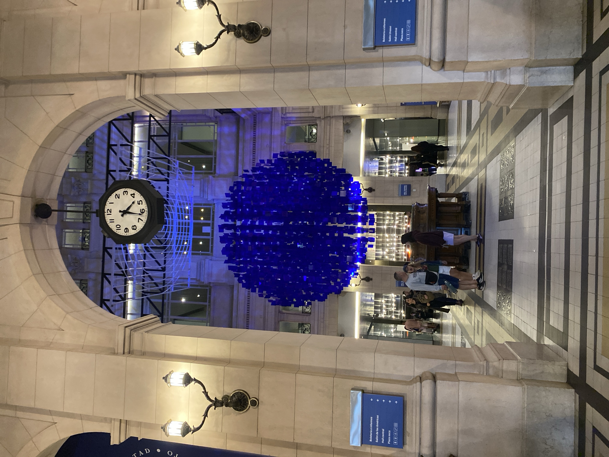 The image shows the interior of a building in Buenos Aires, Argentina. A large clock is visible on the left, reading approximately 5:29. A spherical, blue, illuminated installation hangs in the center of the hall. Several people stand near a reception desk in the middle ground. Two ornate lampposts flank the arched entrance. Signage in Spanish on the columns indicates Boleteria e informes (ticket office and information), Salón de las Américas, Hall Central, and Plaza seca. The floor has alternating dark and light stripes, leading towards the back. The time on the clock is approximately 5:29.