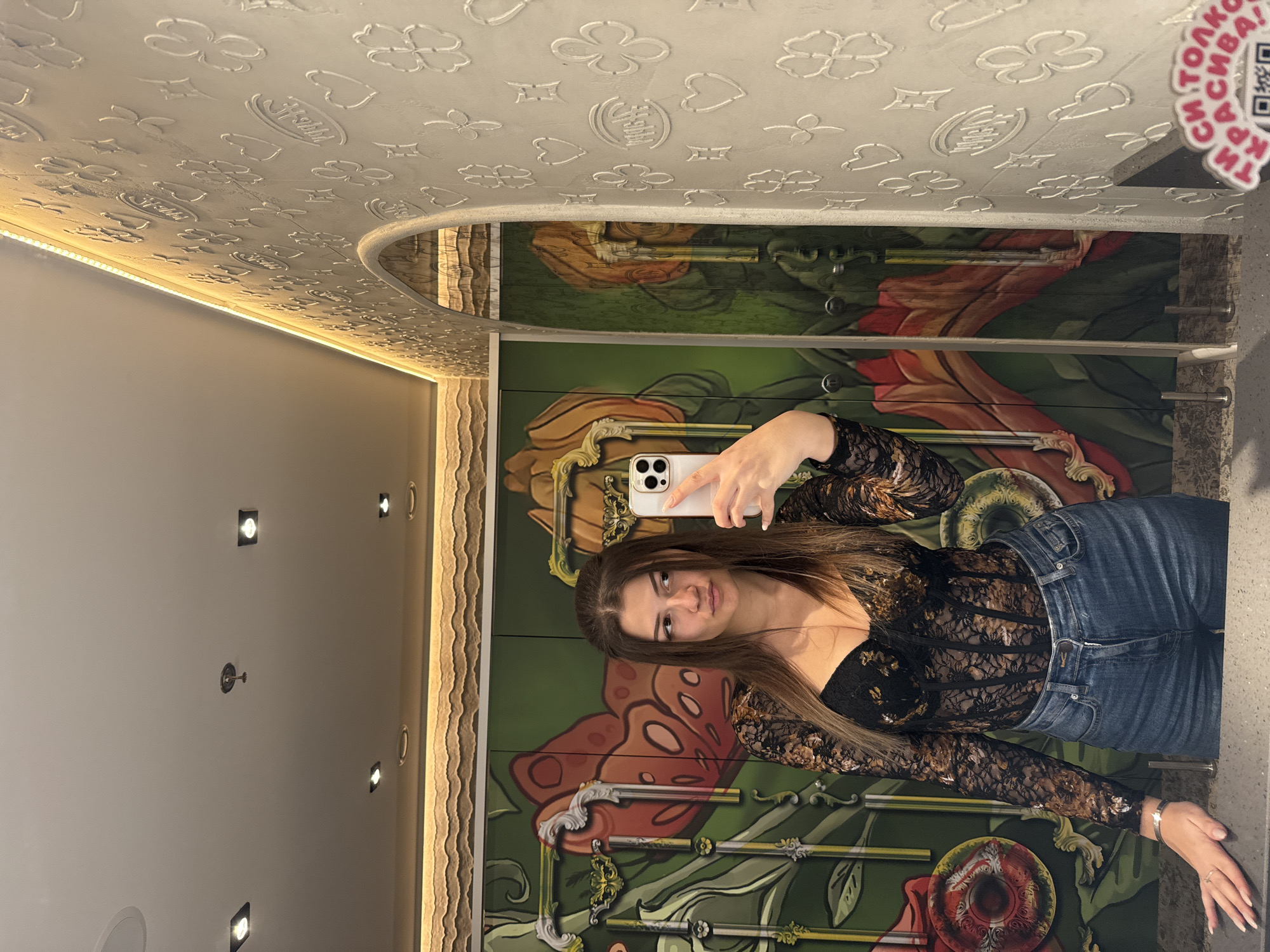 A young adult female is depicted in a mirror selfie within the restroom of Refuel restaurant in Sofia, Bulgaria. She is shown from the chest up, holding a white smartphone with multiple camera lenses in her right hand. Her left hand rests on a dark, mottled countertop that spans the lower portion of the image. She wears a long-sleeved black lace top with gold and bronze floral patterns, and blue denim jeans. A silver bracelet is visible on her left wrist, along with a ring on her left ring finger. Her facial expression is a slight pout, and her gaze is directed towards the viewer's left.
The background behind the subject's reflection features highly decorative, dark green restroom stall doors adorned with oversized orange and red butterfly or insect wings, green foliage, and ornate gold frames. To the right of the mirror, a light-colored wall exhibits embossed patterns of hearts, four-leaf clovers, stars, and a stylized script logo. The ceiling, visible in reflection, is light-colored and fitted with square and circular recessed lights, along with a central fire sprinkler head. A linear LED strip light runs along the upper edge of the embossed wall, providing illumination. On the dark countertop in the lower right, a partially visible pink-and-white circular sticker displays Bulgarian text, "ТИ СИ ТОЛКОВА КРАСИВА!" (YOU ARE SO BEAUTIFUL!), accompanied by a QR code.