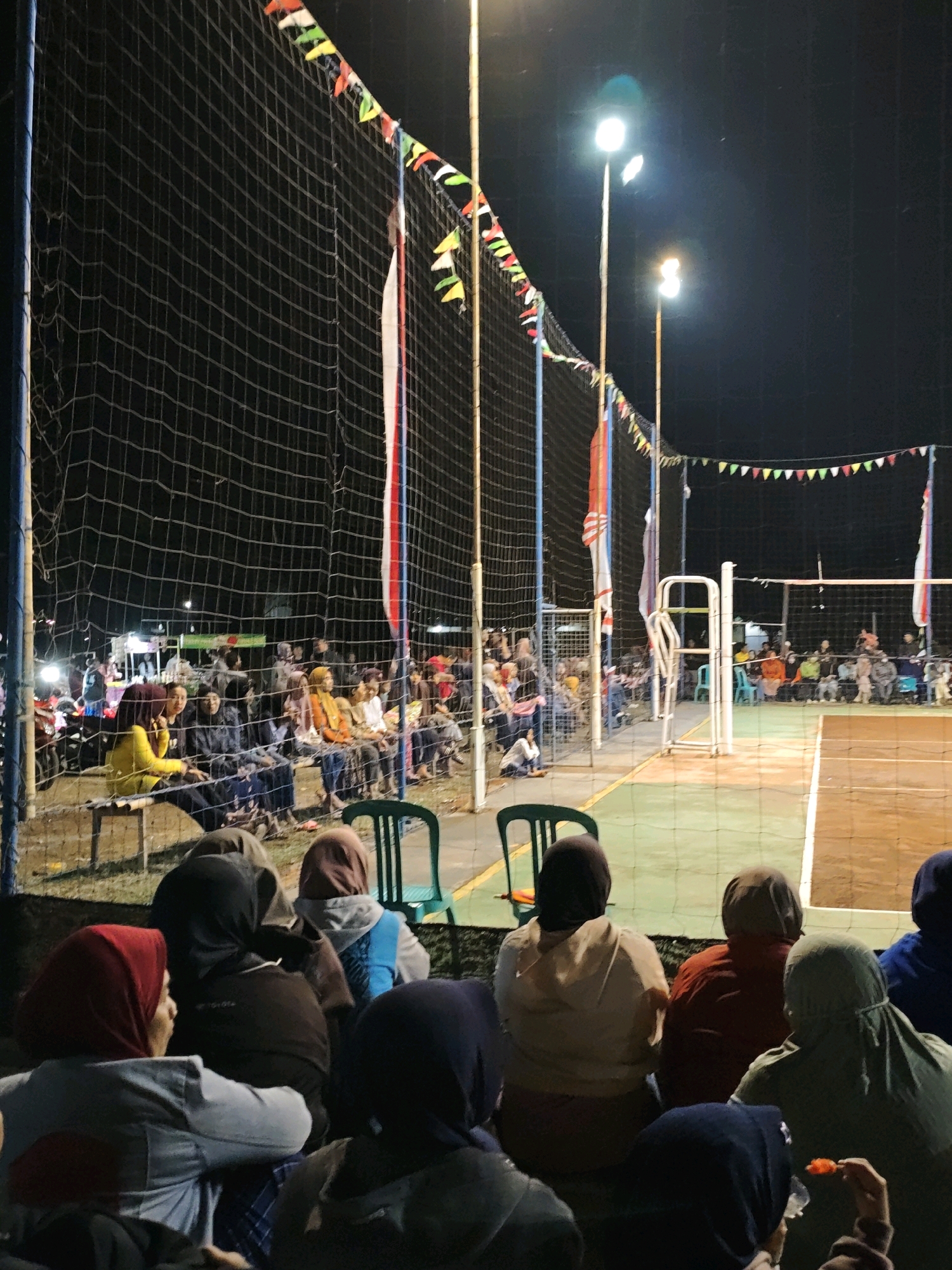 The image depicts an outdoor volleyball game at night in Pagu, Indonesia. A crowd of spectators, many wearing hijabs, are seated on benches behind a volleyball net, watching the game. The court is illuminated by bright floodlights. Decorative flags are strung across the top of the net, adding a festive atmosphere. In the background, more spectators are visible, along with motorcycles and what appears to be a concession stand. The overall scene suggests a community event or local competition taking place in a relatively informal setting.