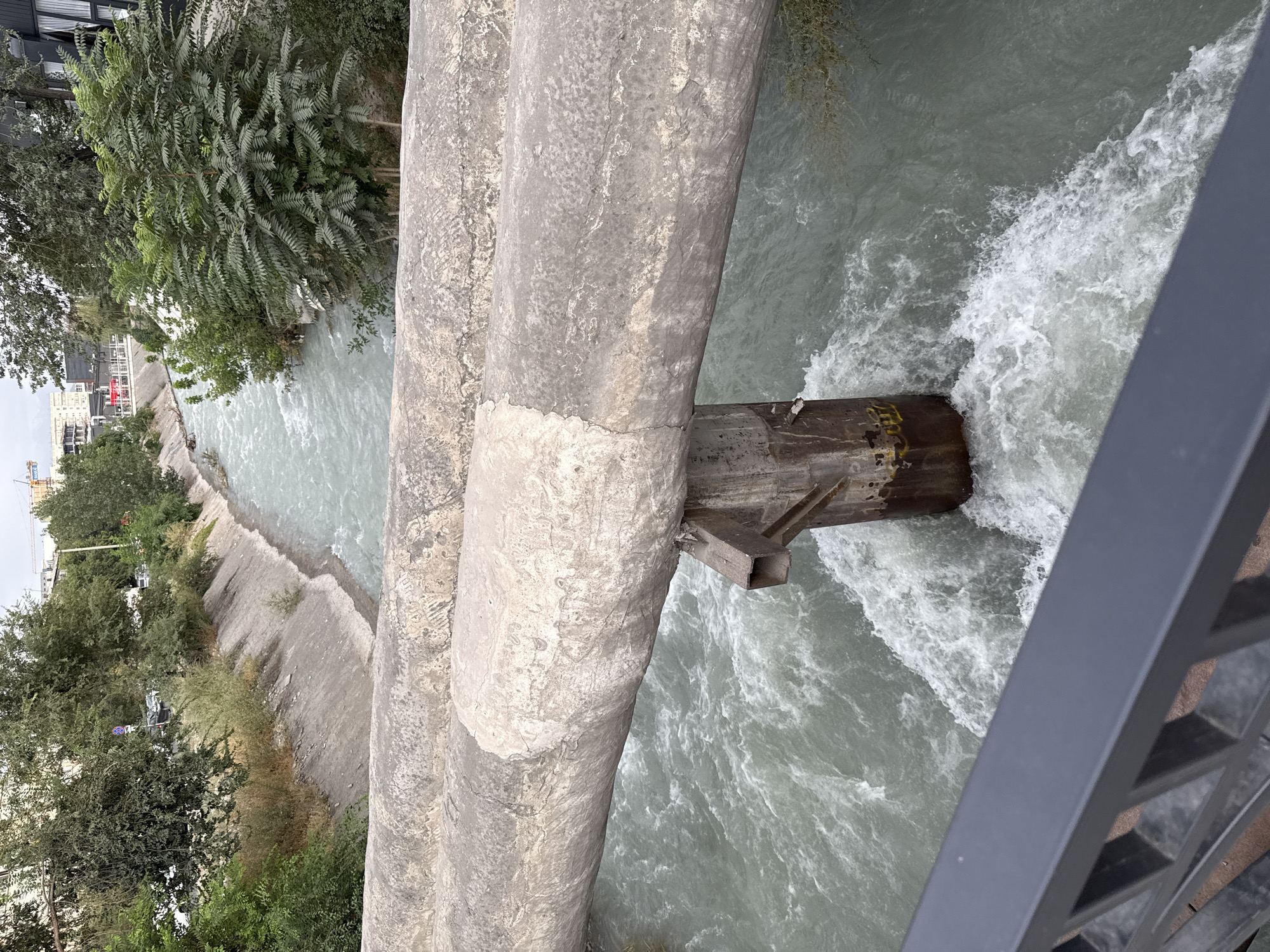 A flowing river runs through a concrete channel with steep banks. On the left bank, there are trees and vegetation, and in the distance, buildings are visible. On the right bank, there are also trees and vegetation, and a building with large windows. Two large concrete pipes are suspended over the river by a concrete support. The water is turbulent and appears to be flowing rapidly. The scene is captured from a slightly elevated viewpoint, possibly from a bridge or walkway, with a metal railing in the foreground. The sky is overcast. This location is identified as Bishkek City, Kyrgyzstan.