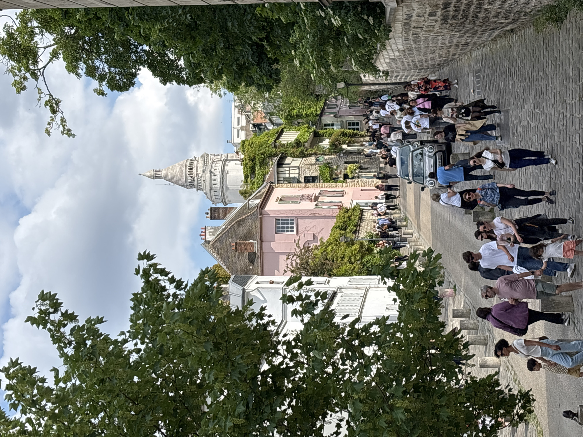 The image captures a vibrant daytime scene on a sloping, cobblestone street, likely in Montmartre, Paris, with the Sacré-Cœur Basilica prominently featured in the background. Numerous people are scattered along the street, appearing to be tourists or locals enjoying a walk. Many are gathered in groups, some standing and observing, while others are walking uphill.
The street is lined with traditional European-style buildings, some with intricate details and a few covered in lush green ivy. A distinctive pink building stands out among the darker stone structures. A small, white retro-style car, resembling a Mini Cooper, is parked or slowly moving amidst the crowd on the right side of the pathway.
Above the buildings and surrounding the street, vibrant green trees with dense foliage provide natural framing. The sky is partly cloudy but bright, indicating a pleasant day. The majestic white dome and spire of the Sacré-Cœur Basilica rise grandly in the distance, against the cloudy backdrop. The scene conveys a lively atmosphere typical of a popular historic and tourist destination. No discernible text is visible in the image.