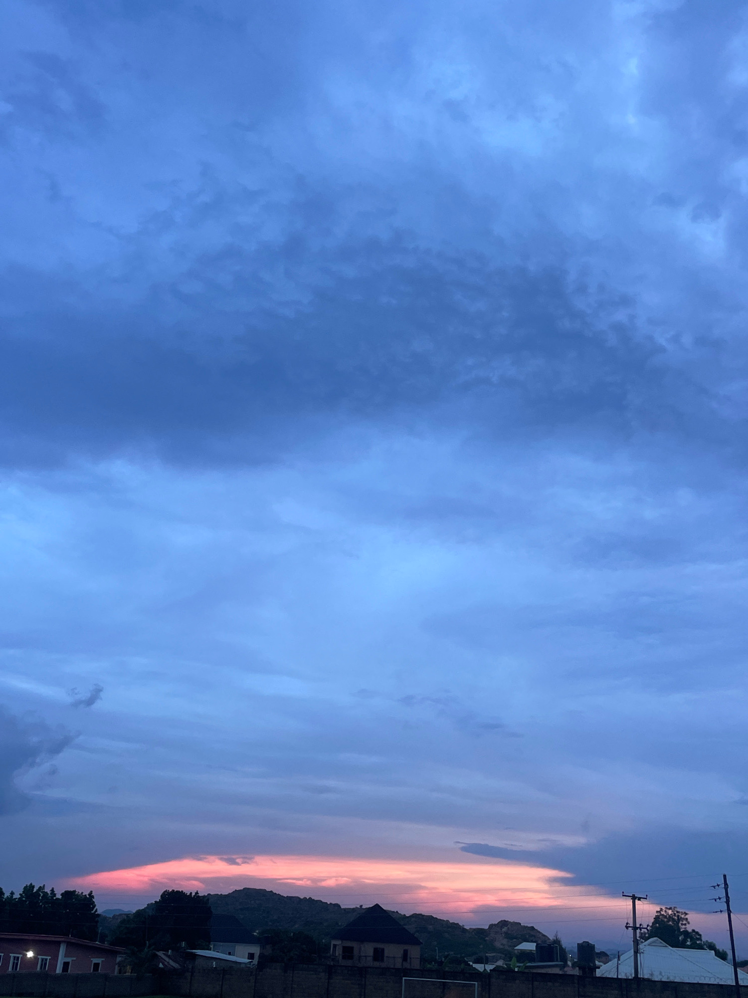 The image depicts a sunset over the city of Jos, Nigeria. The sky is filled with dramatic clouds, transitioning from a deep blue at the top to lighter shades of blue and pink near the horizon. A band of vibrant pink and orange light illuminates the sky just above the silhouette of a distant mountain range. In the foreground, the roofs of several houses are visible, along with trees and utility poles. The overall atmosphere is serene, with the fading light of the day casting a gentle glow over the landscape.