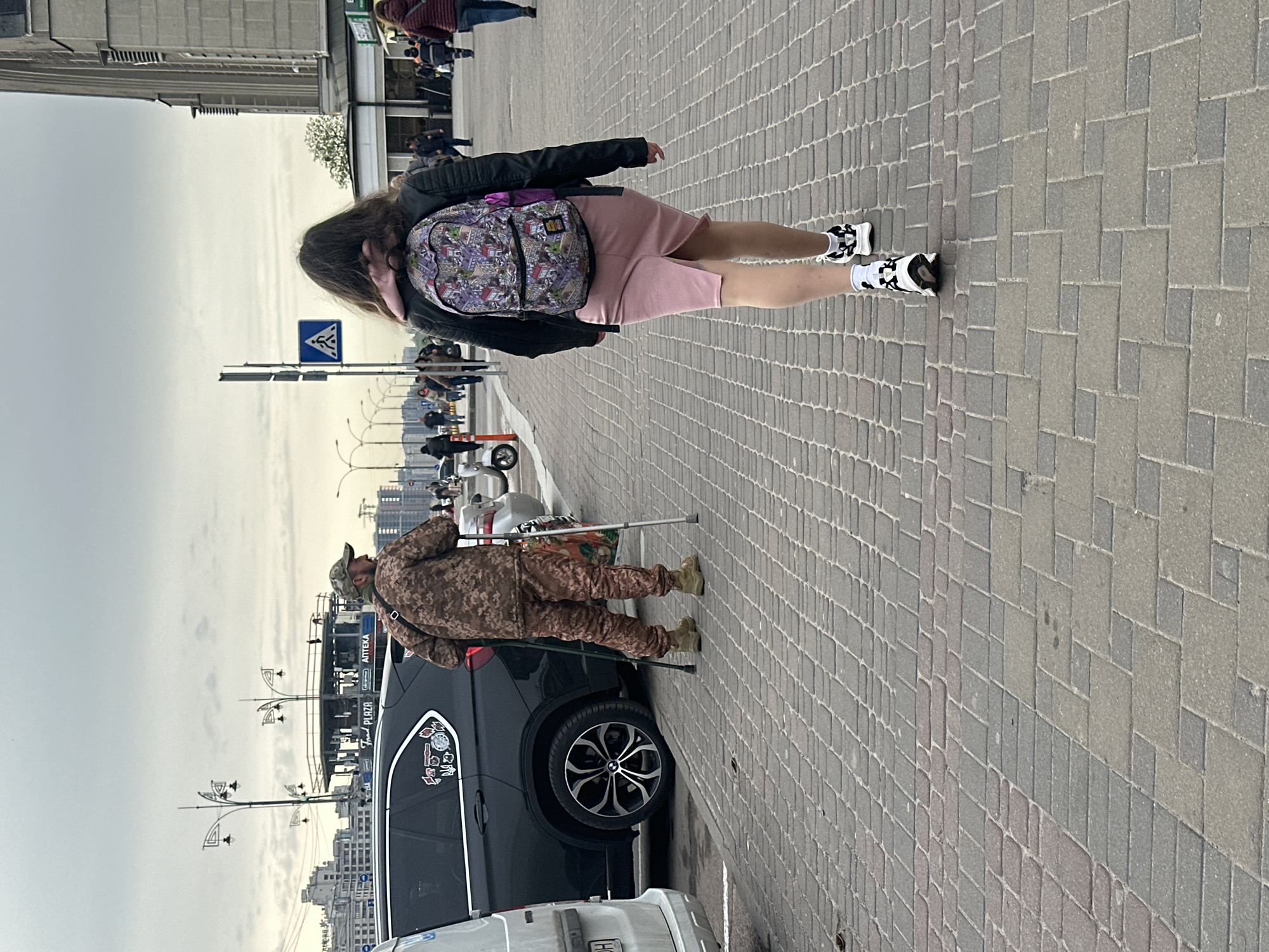 The image captures an urban street scene in Kyiv, Ukraine, under an overcast sky during daytime. The foreground features a woman walking away from the camera on a brick-paved sidewalk. She has long dark hair and is dressed in a black jacket, a pink skirt or dress, and black and white sneakers. A multi-colored, patterned backpack, possibly adorned with cartoon or abstract designs, is on her back.
Further down the street, a man in a camouflage uniform, characterized by a pixelated brown and green pattern, stands near a parked black SUV. He is using crutches, with one visible under his arm. The SUV's rear window displays several stickers, including what appears to be a Ukrainian trident symbol. A partial view of a white vehicle's license plate, showing the letter "H," is visible in front of the SUV.
The background includes various urban elements: tall buildings, streetlights, and traffic signs, one of which is a blue square sign with a white arrow. A building in the distance prominently features text signs for "PLAZA" and "APTEKA" (pharmacy). The sky is uniformly light grey, indicating an overcast day. The street and pedestrian areas are paved with rectangular grey pavers of different shades.