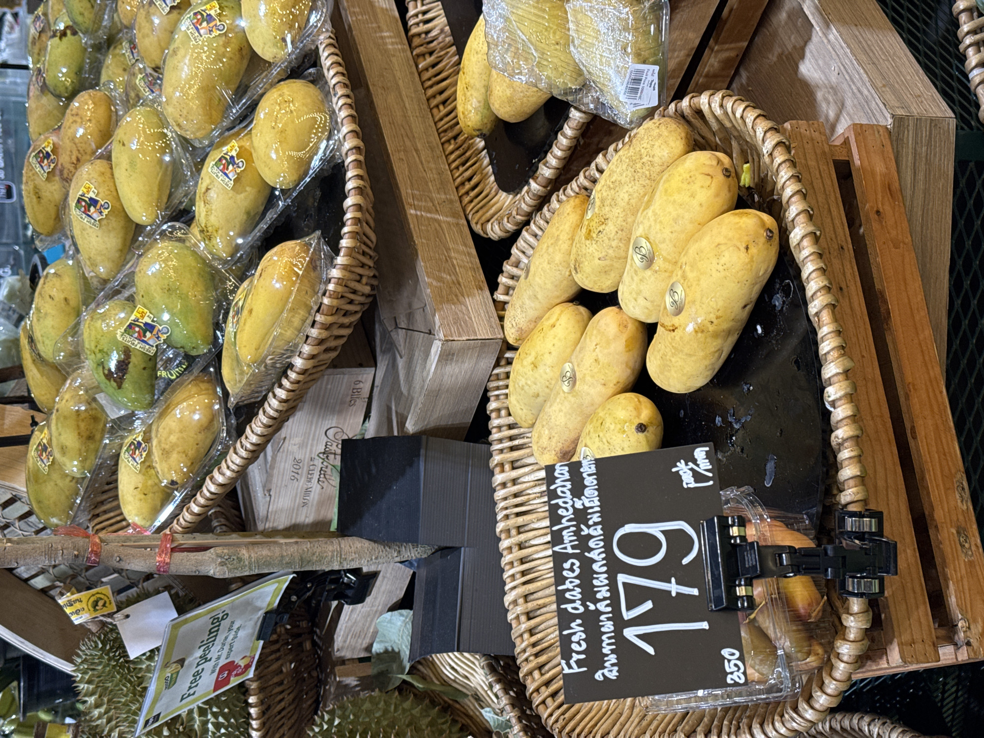 The image depicts a fruit stall in the city of Ko Samui District, Thailand. The foreground features a basket filled with yellow mangoes, some of which are packaged in plastic. A black sign with white lettering displays "Fresh dabs Amhedahan" and a price of "179". To the left of the sign, a smaller price of "350" is visible. Behind the mangoes, durian fruits are partially visible. In the background, several rows of mangoes, also packaged in plastic, are stacked on shelves, with various signs and labels associated with the fruits. The overall environment suggests a market setting with an abundance of fresh produce.