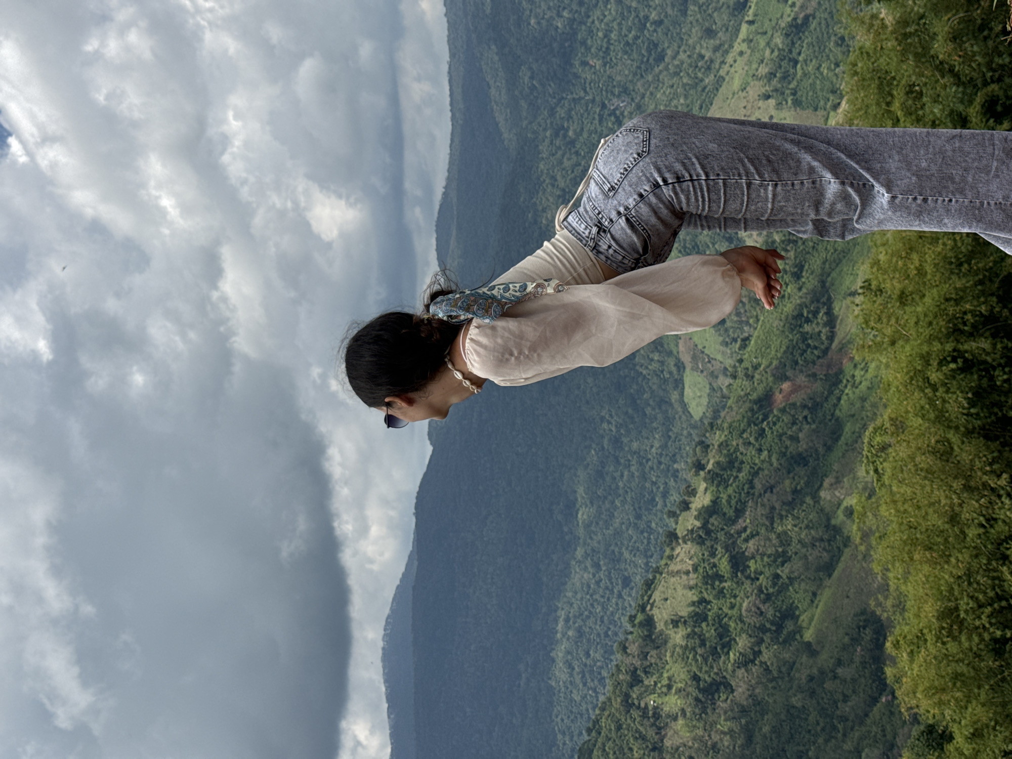 A person, viewed from the side and from the back, is leaning forward to look out over a vast, mountainous landscape in Nao Sẻ, Vietnam. The person has dark hair tied back with a blue and white patterned scarf and wears sunglasses, a sheer, light-colored blouse with billowy sleeves, and light-wash gray jeans. The blouse is layered over a beige tank top, and a pearl necklace is visible around the person's neck. The person's hands are clasped together in front.
The background consists of rolling hills covered in dense green forest, receding into layered, misty mountains under a cloudy, gray sky. In the foreground, lush green foliage, including bamboo, forms a dense border along the bottom edge of the frame. The terrain appears to be a scenic overlook or viewpoint.