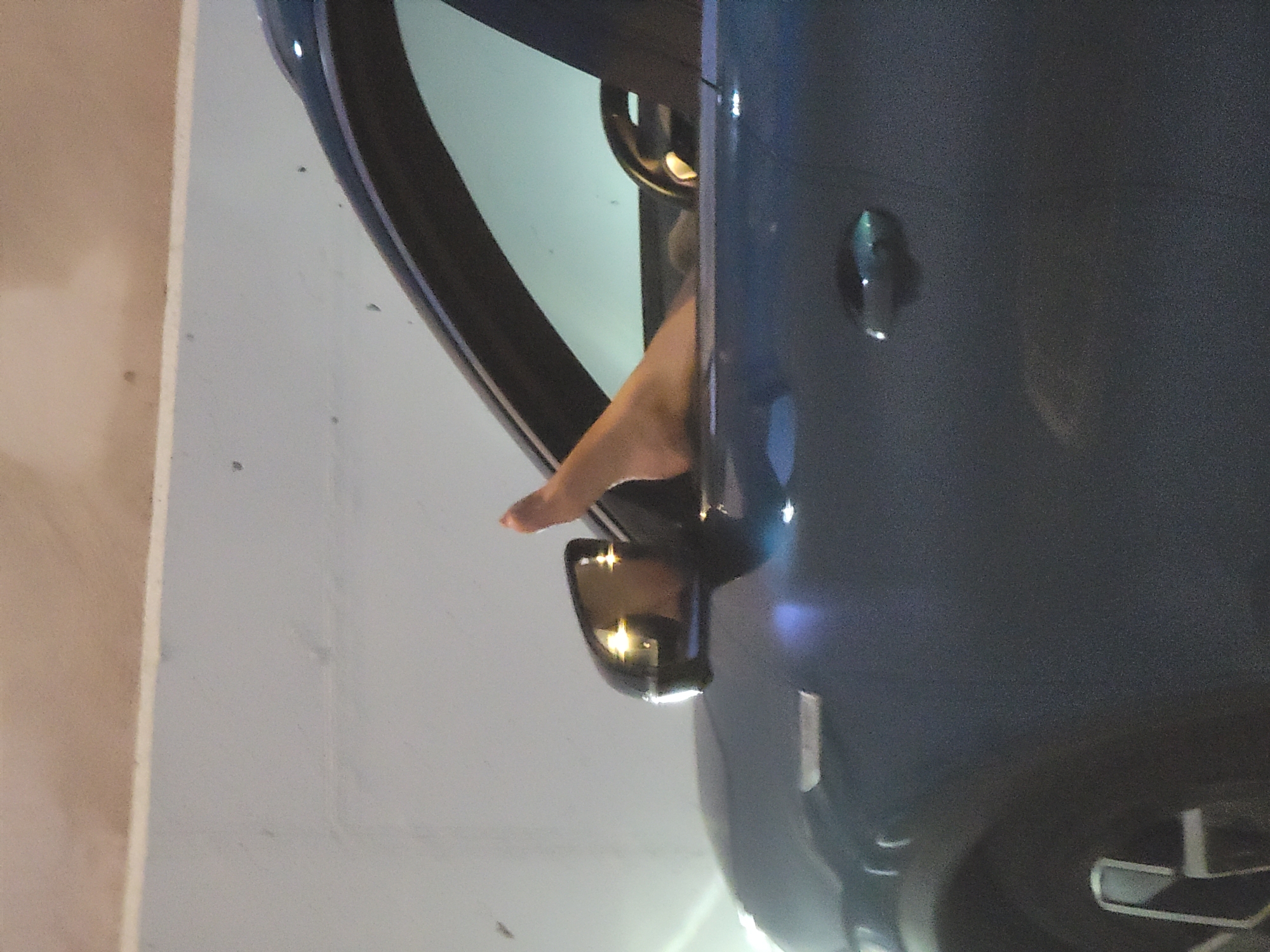 A person's bare foot is visible protruding from the open driver's side window of a dark blue car. The car is parked outdoors, with a concrete wall in the background. The foot appears to be relaxed, with the toes pointing upwards. The interior of the car, including a steering wheel, is partially visible through the window. The lighting suggests it may be dusk or nighttime. The image provides no information regarding the specific location in Quarteira, Portugal.
