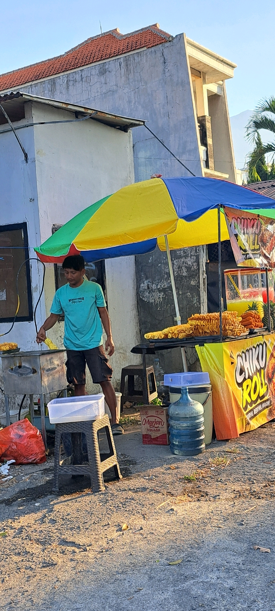 This is a vertical, eye-level, medium shot of a food vendor in Prigen, Indonesia, in front of a light blue building. The vendor, a young man wearing a teal t-shirt and dark shorts, stands to the left of a food cart and appears to be preparing food. He is holding a utensil over a stainless steel fryer or cooker. The food cart is covered by a multi-colored umbrella, with sections of yellow, blue, and green. The cart itself has a yellow front panel with the text "CHIKU ROLL" prominently displayed. Behind the vendor and to the right of the cart, a large blue water jug is visible, along with a cardboard box and some plastic containers. The ground is paved and appears slightly worn, with some debris scattered around. The background includes the side of the building, which has a weathered facade and a red-tiled roof. A palm tree is visible in the upper right corner. The lighting suggests it is daytime, possibly late afternoon given the long shadows.