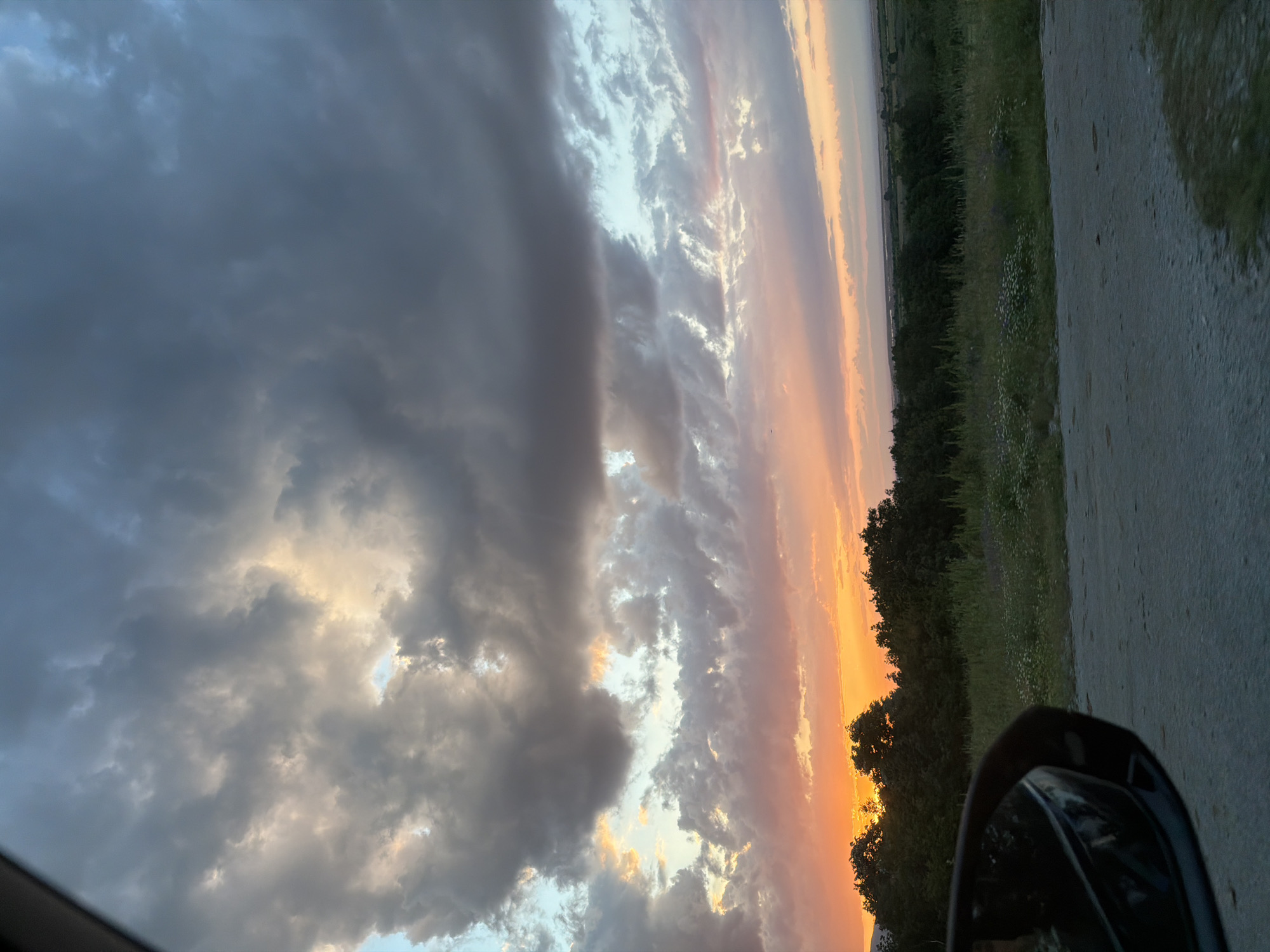 The image, taken from inside a vehicle, shows a vivid sunset in Novakovo, Bulgaria. Dense, dark clouds fill the left portion of the frame, while a strip of bright orange and pink hues illuminates the sky to the right. Below the horizon, there is a treeline and greenery. A section of asphalt road and the vehicle's side mirror are visible in the foreground on the right.