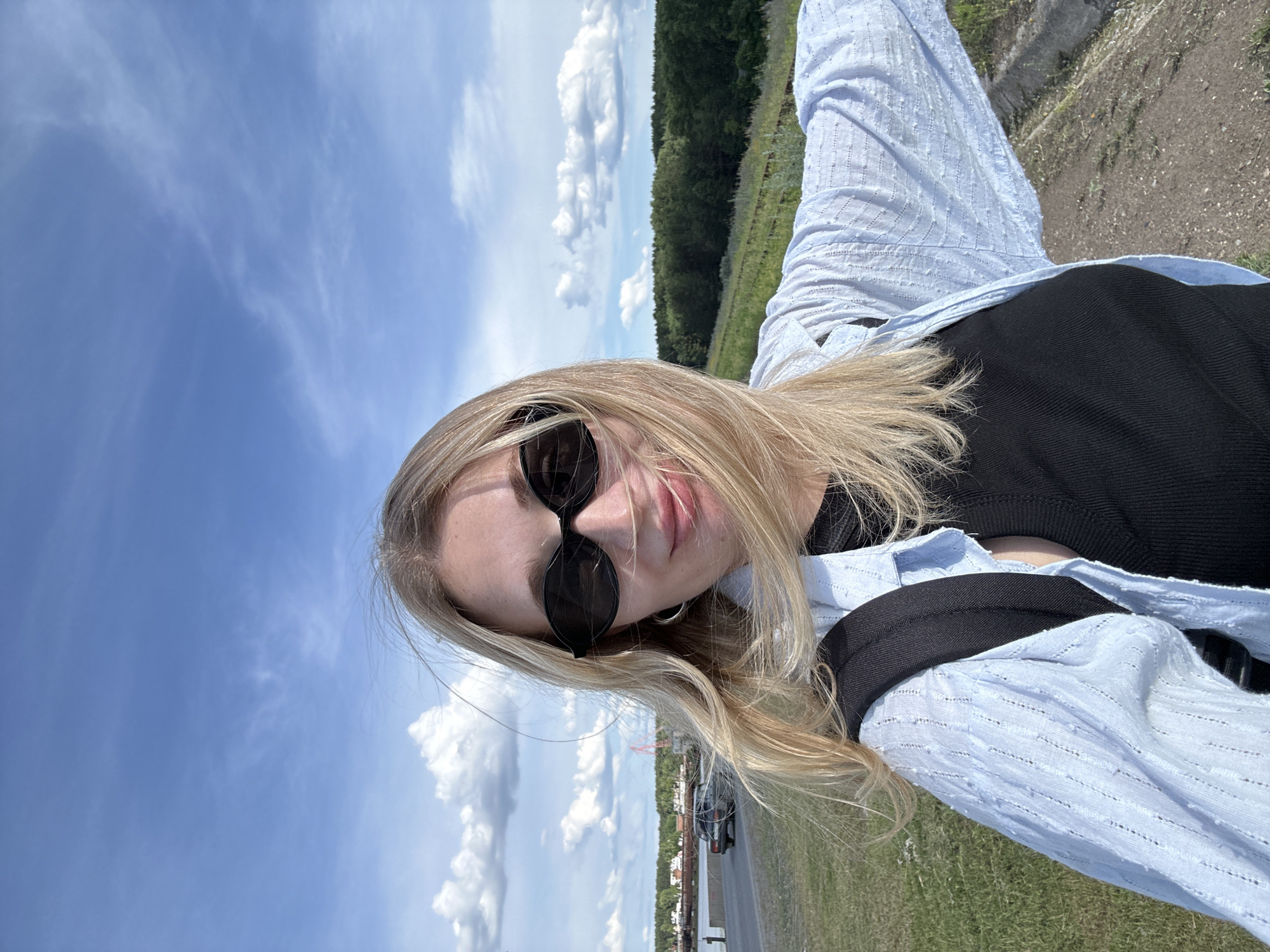 The media file is a portrait-oriented, low-angle selfie of a young Caucasian woman with blonde hair, wearing sunglasses and a light blue collared shirt over a black top. Her hair is long and wavy, framing her face. She is outdoors on a sunny day with a clear blue sky filled with fluffy white clouds.
In the background, a line of trees forms a distant horizon, with a field of grass and some gravel in the foreground. Further in the distance, a road is visible with a dark-colored car. There are also some structures that appear to be buildings and possibly a red industrial structure on the left side of the frame. The overall setting suggests an outdoor, potentially rural or semi-rural location.
The lighting indicates it is daytime, likely midday given the brightness of the sun. The weather appears to be warm and pleasant. The woman's expression is neutral or slightly smiling, with her lips slightly parted. The image conveys a casual, relaxed mood. There is no visible text in the media file.