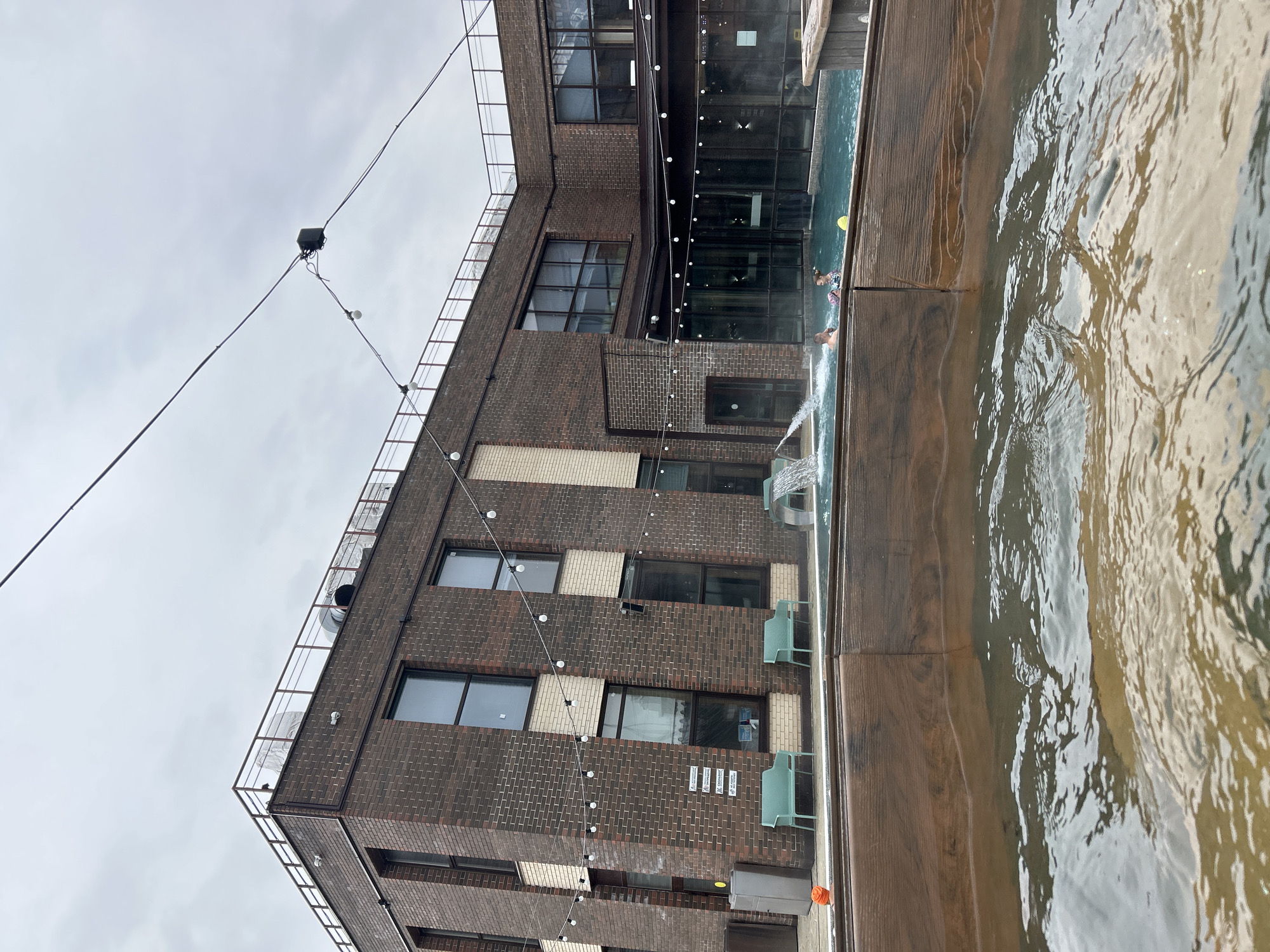 An outdoor scene in Tver, Russia, features a multi-story building constructed primarily of dark brown and light beige bricks, with numerous large windows. The sky above is overcast and light grey, suggesting a cloudy day.
Adjacent to the building is a large swimming pool or hot tub, edged with dark wooden decking. Steam appears to be rising from the water, indicating it might be heated, which, coupled with what looks like small patches of snow or ice on the building's roof railing, hints at a colder climate. Several people are visible in the pool, partially submerged, engaging in recreational activity. A small waterfall feature from one section of the pool cascades into another.
Overhead, a series of string lights with exposed bulbs stretch across the scene, likely for evening illumination. A black box-like object is suspended from the wires. On a lower balcony or ledge of the building, two empty green chairs are positioned.
Visible text on the building's brick wall includes a white sign with the word "КУРЕНИЕ" (Smoking), likely part of a no-smoking directive. Below it, another sign clearly states "ОБЬЕКТ НАХОДИТСЯ ПОД ВИДЕОНАБЛЮДЕНИЕМ" (Object is under video surveillance). The scene conveys a calm, recreational atmosphere during daytime.