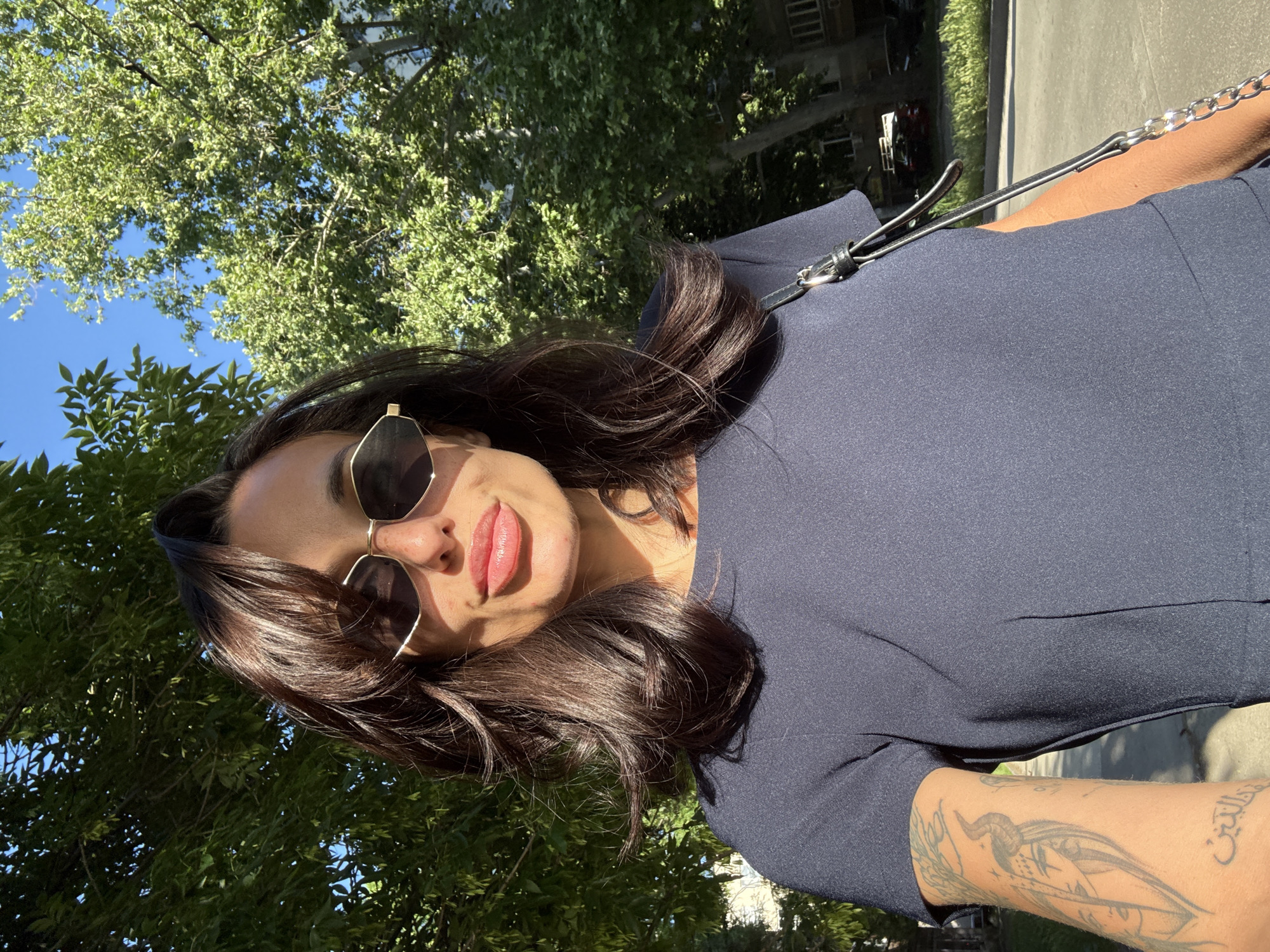 The image shows a woman with sunglasses and a dark blue shirt, outdoors. The setting appears to be Pleven, Bulgaria, with green trees and an urban background, possibly near a street or park. The woman's arm has a tattoo visible. She appears to be holding the camera or phone to take a selfie.