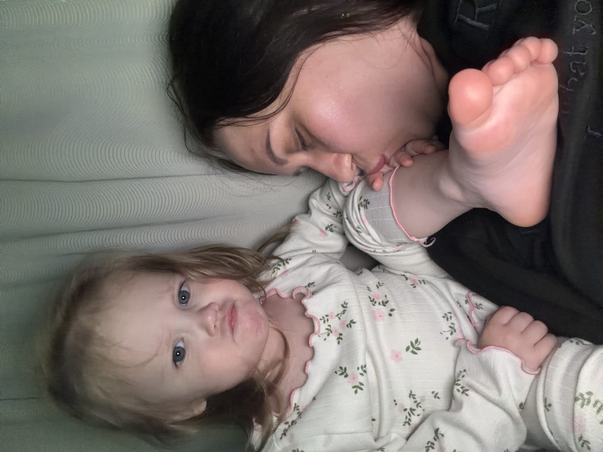 In a close-up, indoor shot, a toddler with blonde hair and blue eyes, wearing white pajamas with a floral pattern, makes a pouting face. Next to the toddler, a woman with dark hair is seen kissing the toddler's foot. The background consists of a light-colored, textured curtain. The image does not provide any information about the city of Perm, Russia.