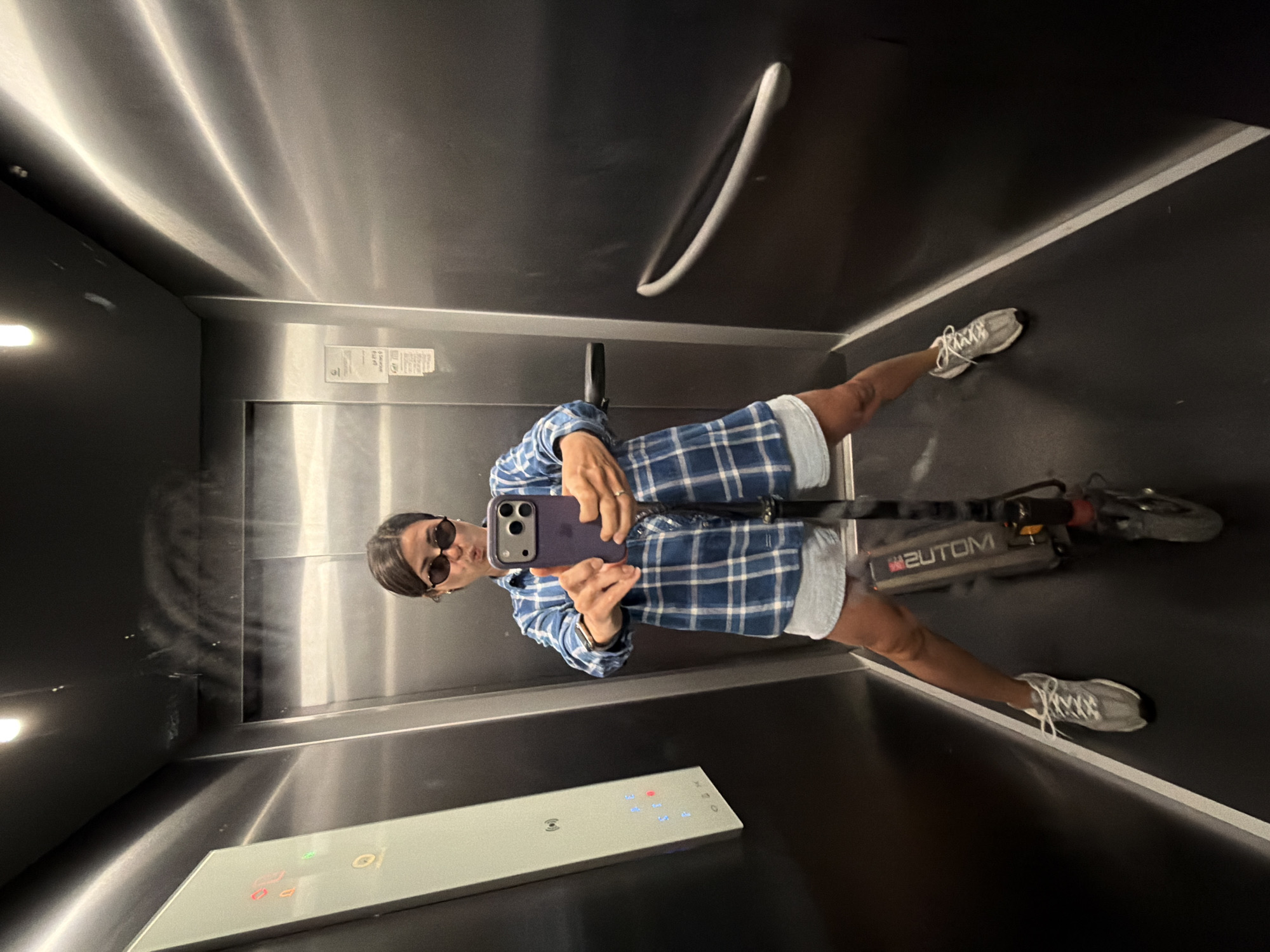 A woman in sunglasses and a blue plaid shirt stands inside an elevator, holding a purple iPhone in front of her to take a selfie. She wears grey shorts and white sneakers, and a scooter is positioned on the floor next to her. The elevator has stainless steel walls and black floor. A control panel with buttons is visible. The elevator is located in the city of Budva, Montenegro, as evidenced by the "2UTOM" logo on the scooter, a local company.