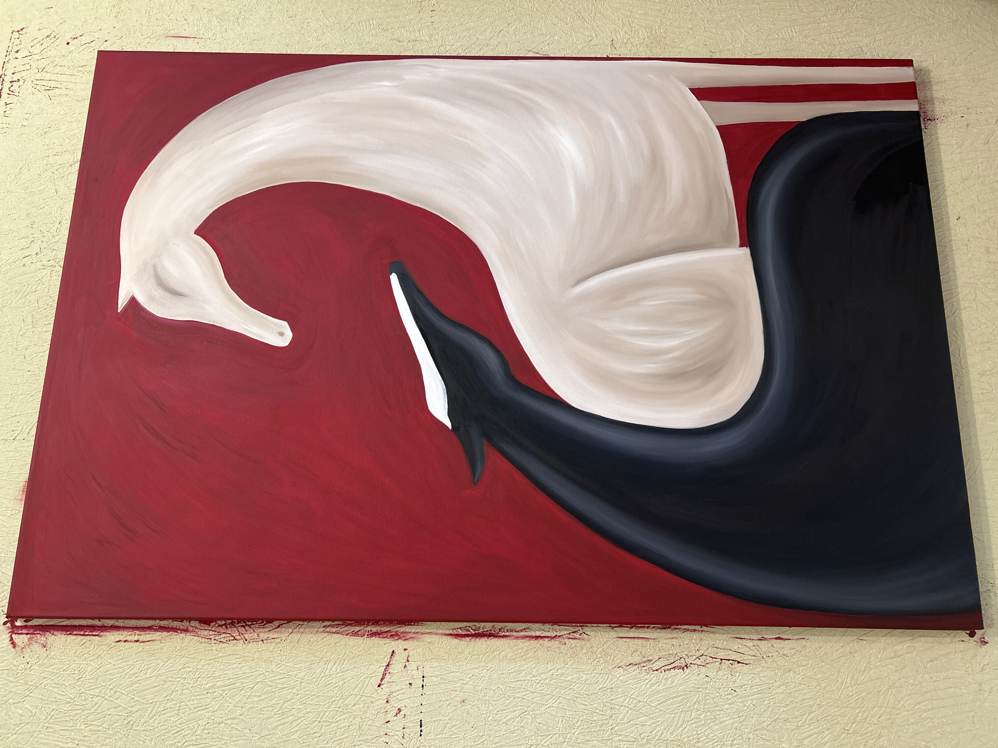 This is a vertical, close-up, eye-level photograph of a painting leaning against a textured, cream-colored wall. The painting, rendered in acrylics, depicts two stylized horse-like figures against a solid red background. The figure on the right is white with subtle shading, its head and neck curved in a graceful arc. The figure on the left is primarily black with a white stripe across its head and neck, and its form is curved and appears to be intertwined with or facing the white figure. The canvas edges of the painting show red paint drips and smudges, indicating it is likely still wet or has been recently handled. The wall texture is rough, with visible vertical lines and a few red paint marks. The overall impression is one of elegance and contrast, with the bold red background and the opposing colors of the figures. There is no indication of its location in Moscow, Russia.