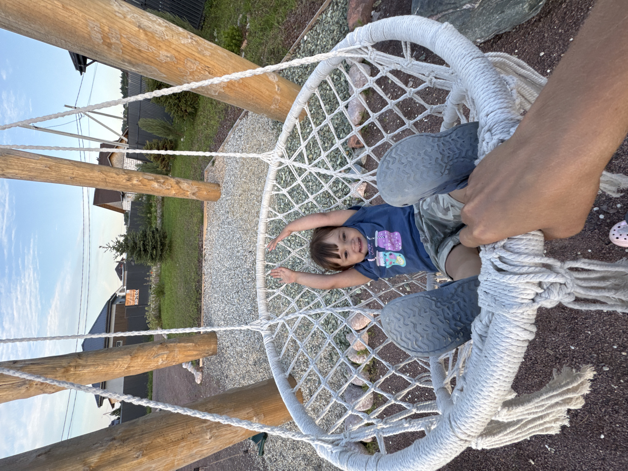 In Kempelevo, Russia, a young child with dark hair and a blue t-shirt is sitting in a white macrame hanging chair suspended from wooden posts. The child is smiling and has their arms raised. They are wearing camouflage shorts and grey slip-on shoes. The chair is positioned outdoors on a gravelly surface, with a green lawn and residential buildings visible in the background. The sky is blue with some clouds. A person's hand is visible in the foreground, holding onto the chair.